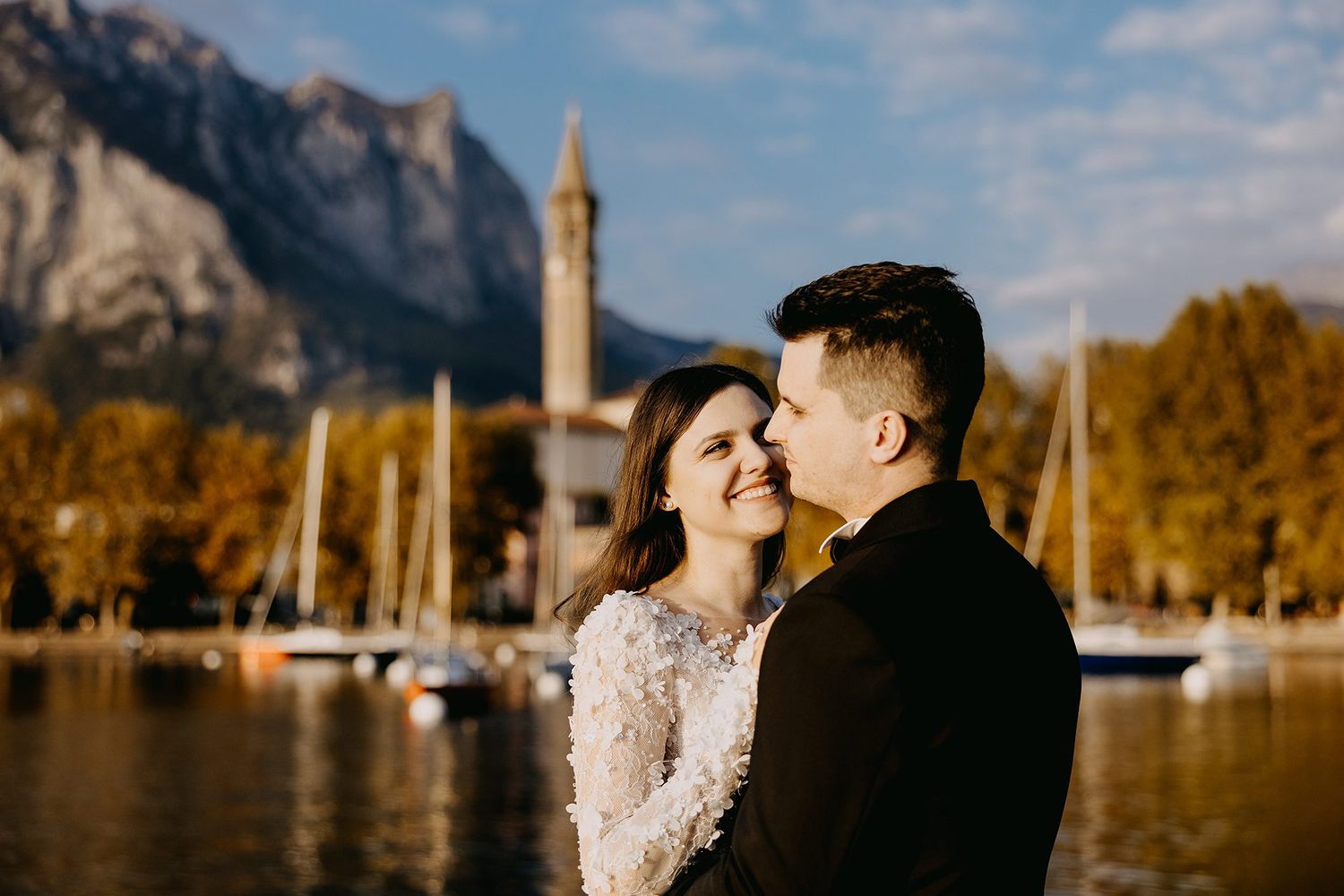 Smiling bride and groom share a tender moment by a lakeside marina with mountain views.