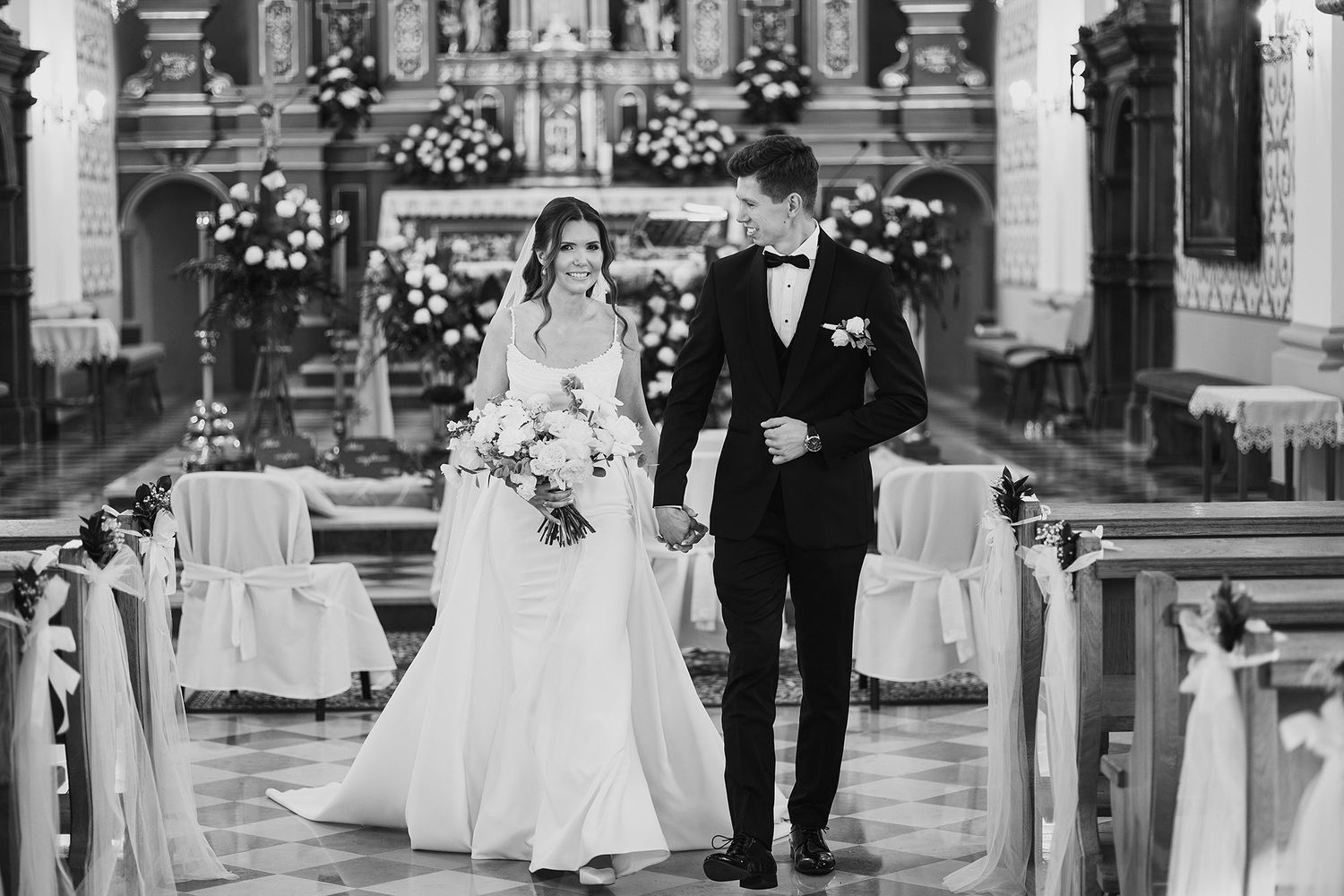 Black and white portrait of bride and groom walking hand in hand down a decorated church aisle.