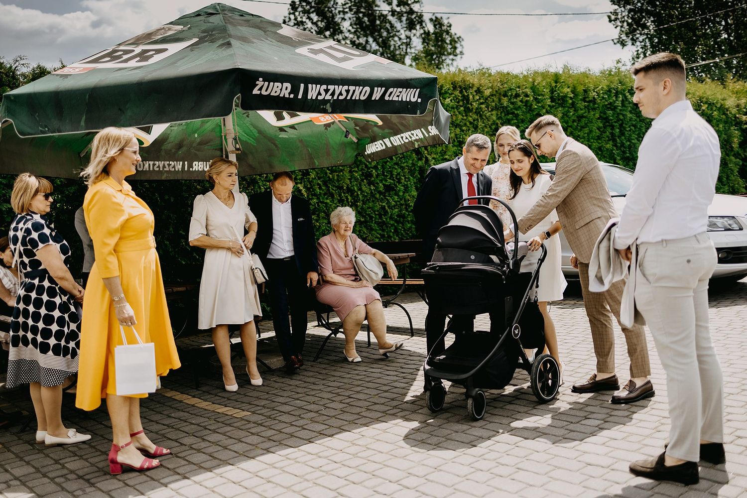 Family group gathering outdoors near a stroller after a christening ceremony on a sunny day.
