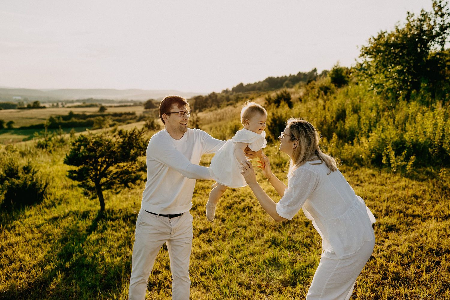 Happy young family in white outfits playing with their baby in a sunny green meadow at sunset.
