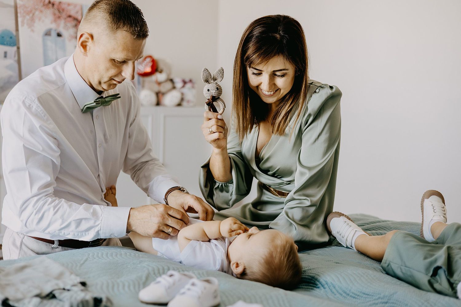 Parents in elegant attire playing with baby using bunny toy during baptism day preparations.