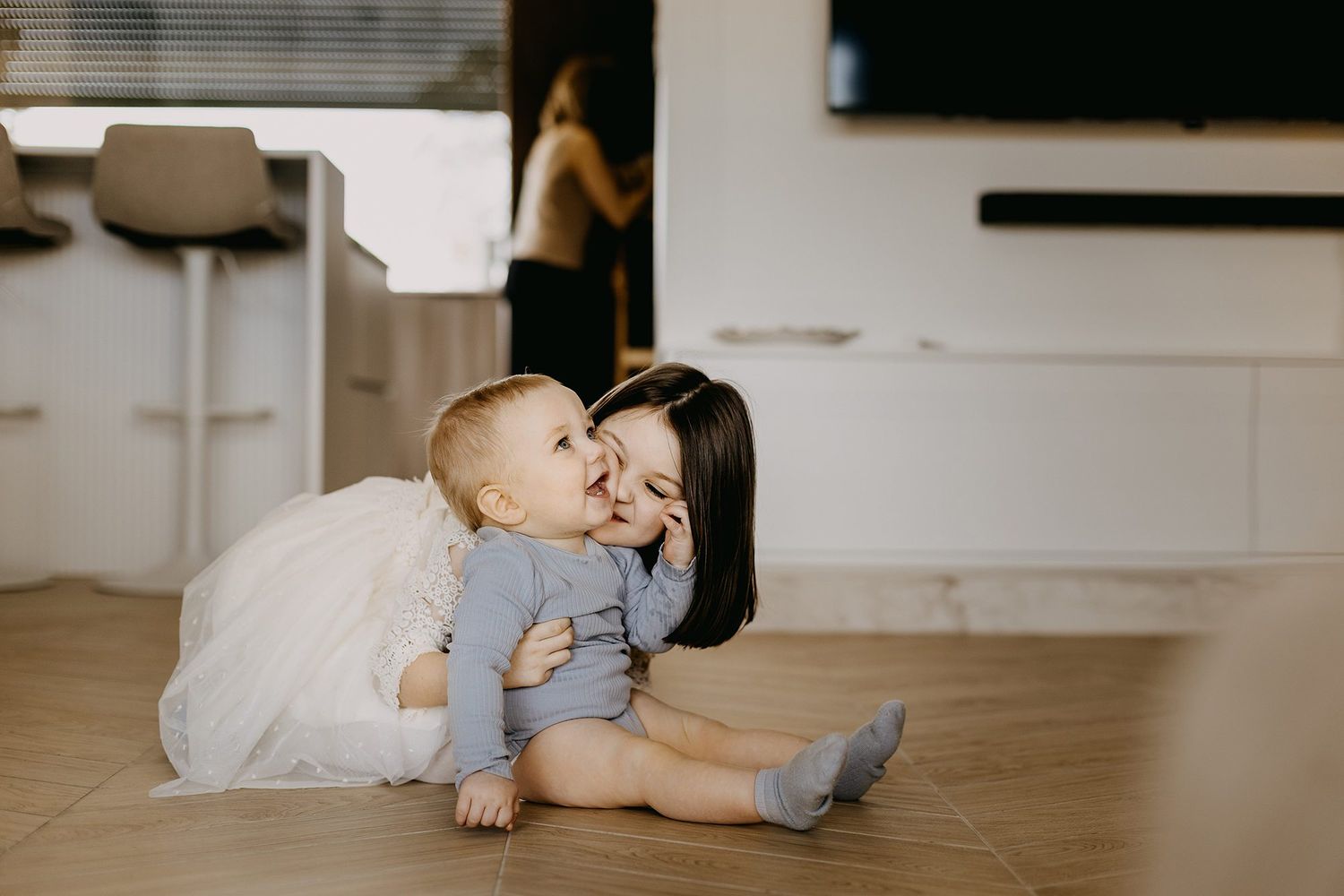 A young girl kisses a smiling baby sibling while sitting on a home floor.