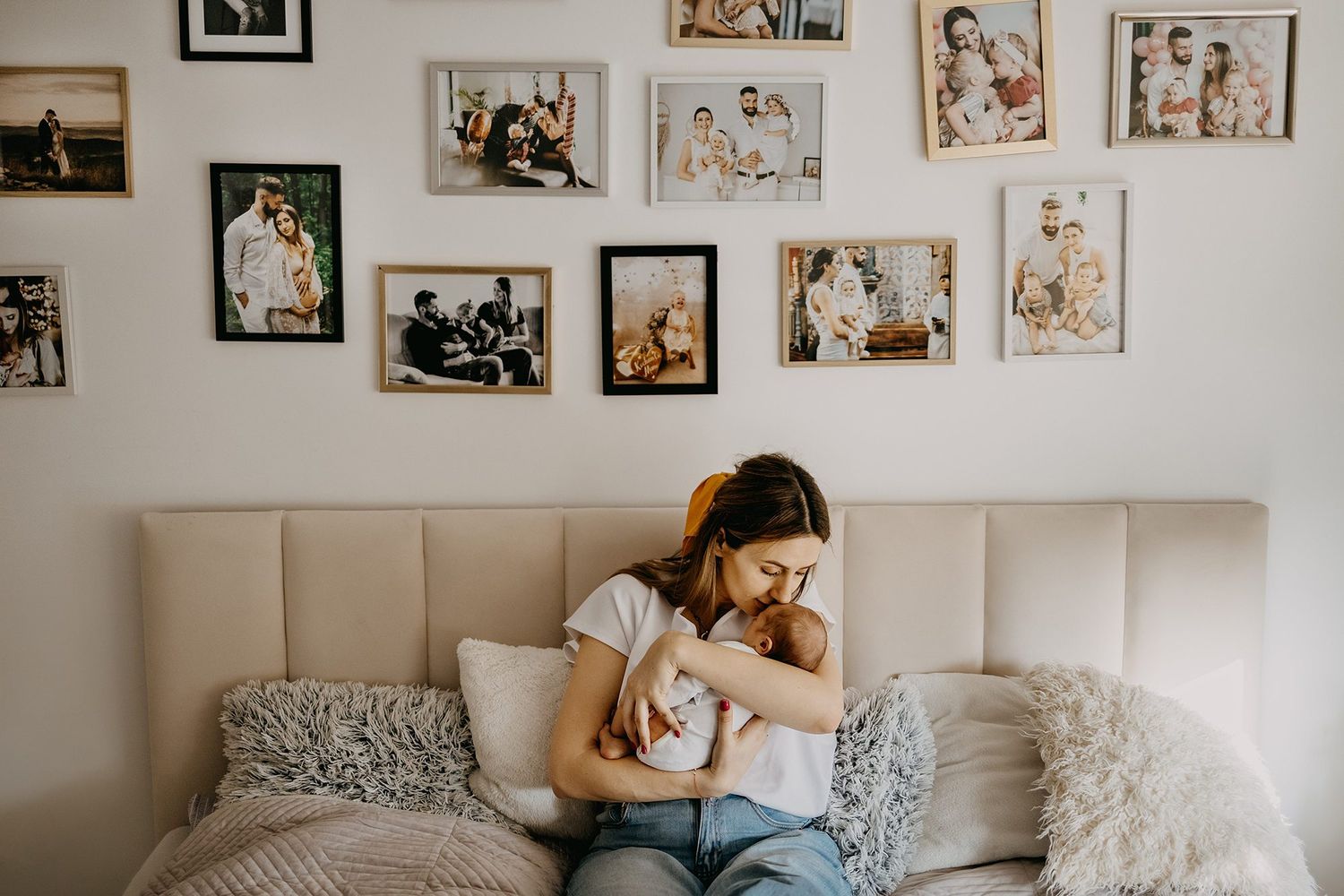 A young mother sits on a bed holding her newborn, surrounded by framed family photos.