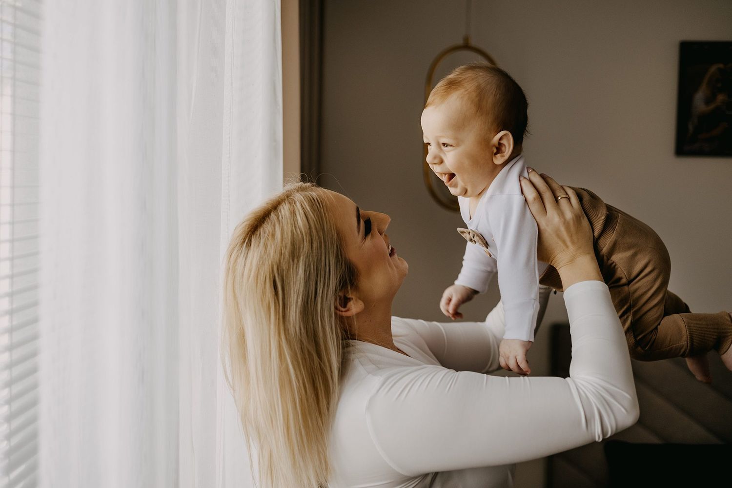 Joyful blonde mother lifting her laughing baby near a bright window in a cozy home setting.