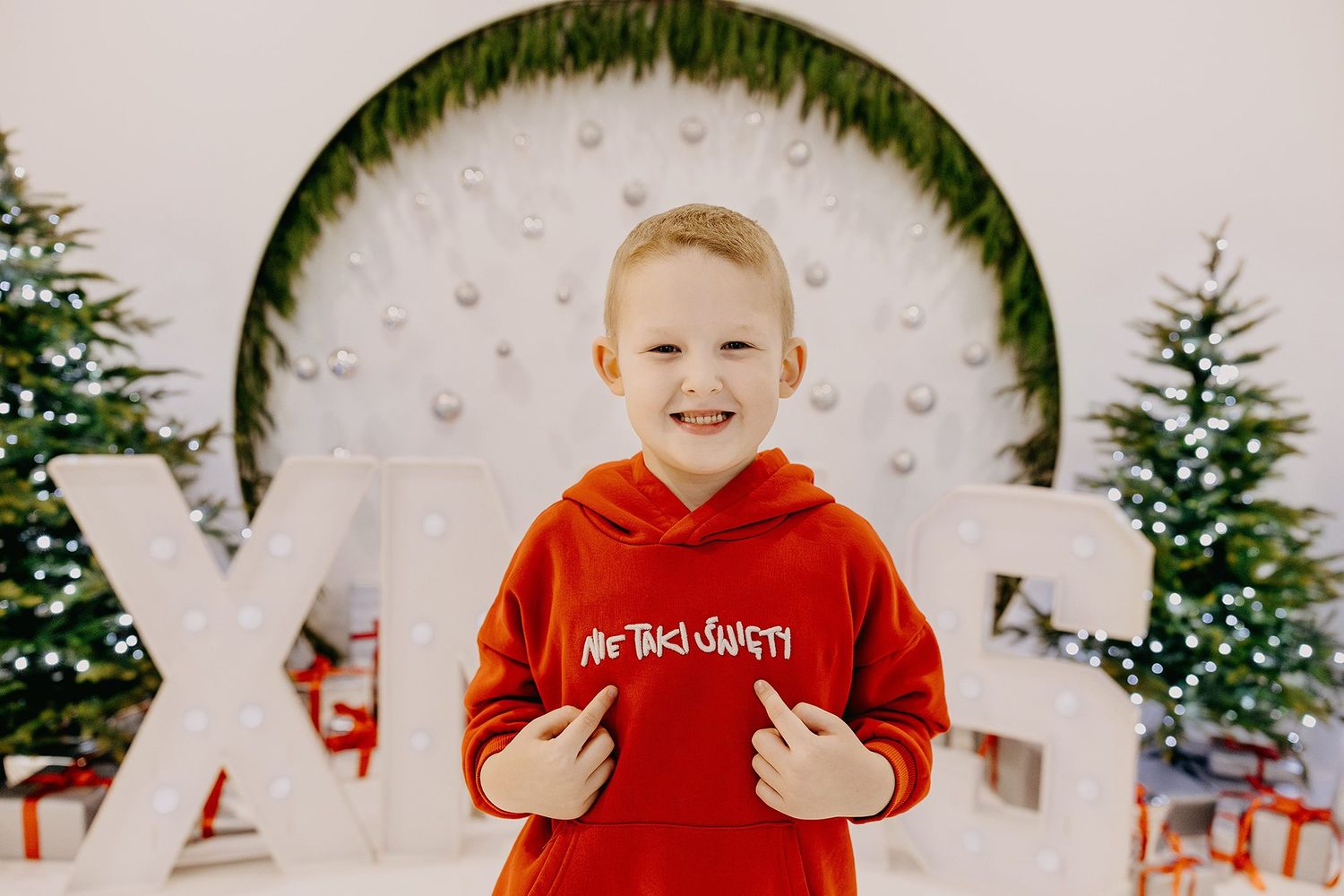 Smiling young boy in red hoodie posing in front of Christmas trees and festive holiday decorations.