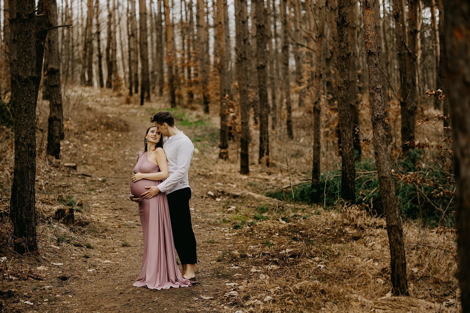 Romantic couple in pine forest, man kissing pregnant womans head as she holds her pink gown.