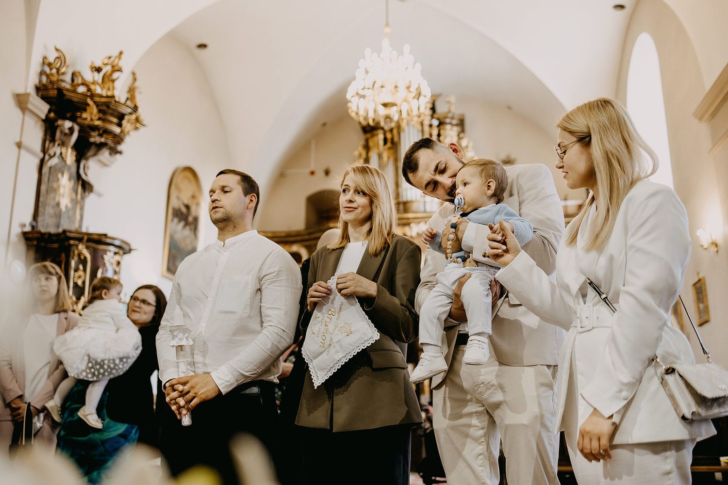 Family group holds baby during christening ceremony inside ornate vaulted church interior.