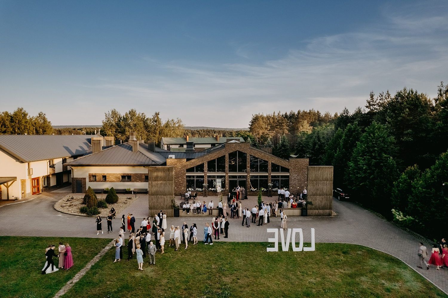 Aerial view of a wedding venue with guests gathered outside a rustic building, featuring a large LOVE sign at golden hour.