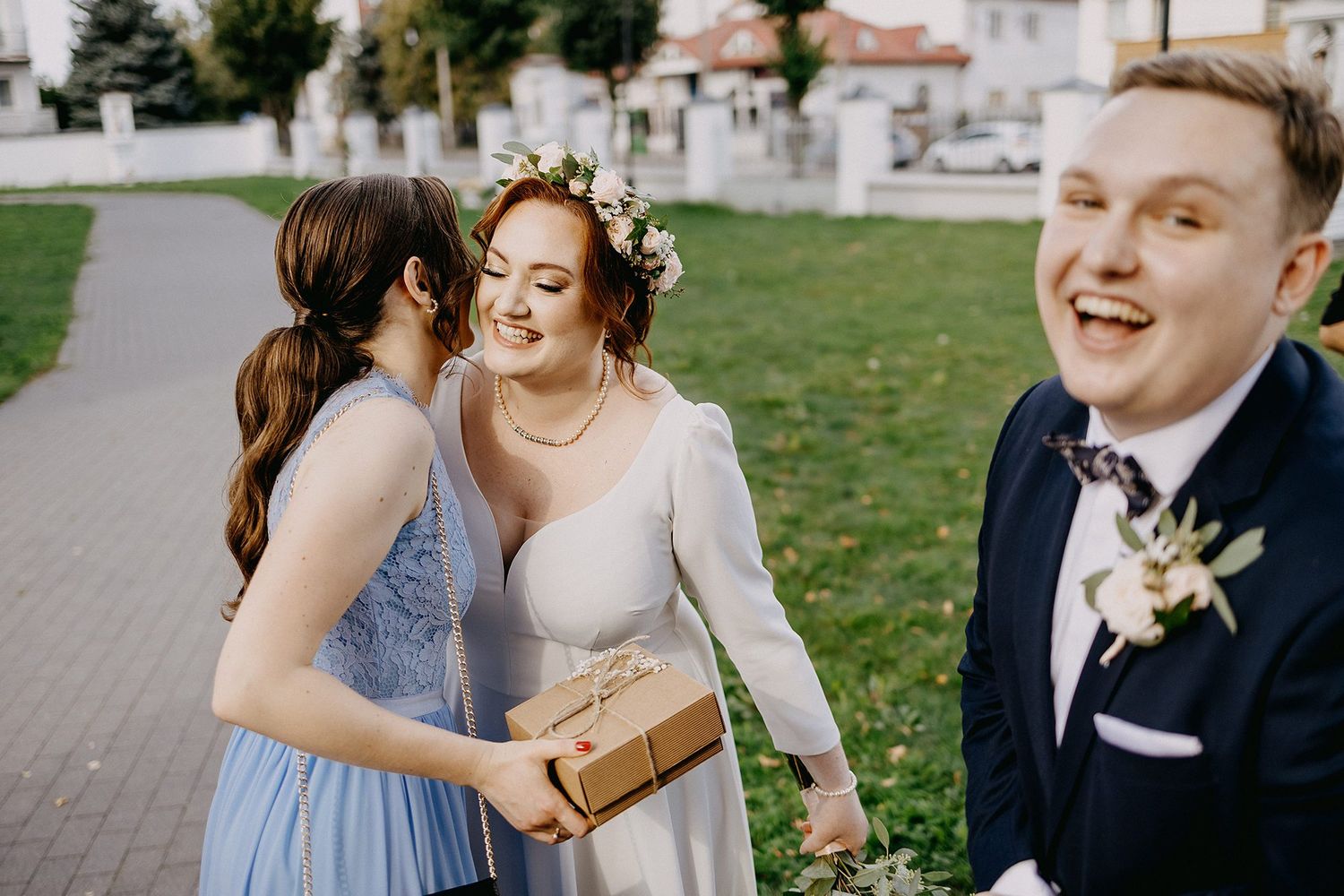 Bride in white dress with floral crown receives gift from bridesmaid in blue, as groom laughs joyfully outdoors.