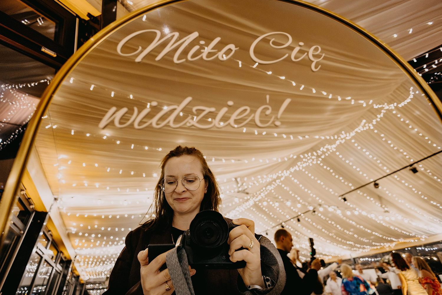 Woman using smartphone under elegant wedding tent with fairy lights and cursive Polish welcome sign.