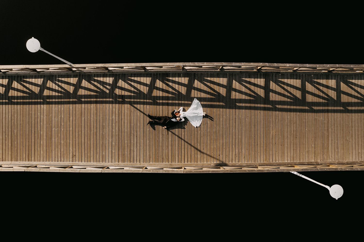 Aerial drone view of bride and groom walking on a wooden bridge, casting dramatic shadows on a sunny day.