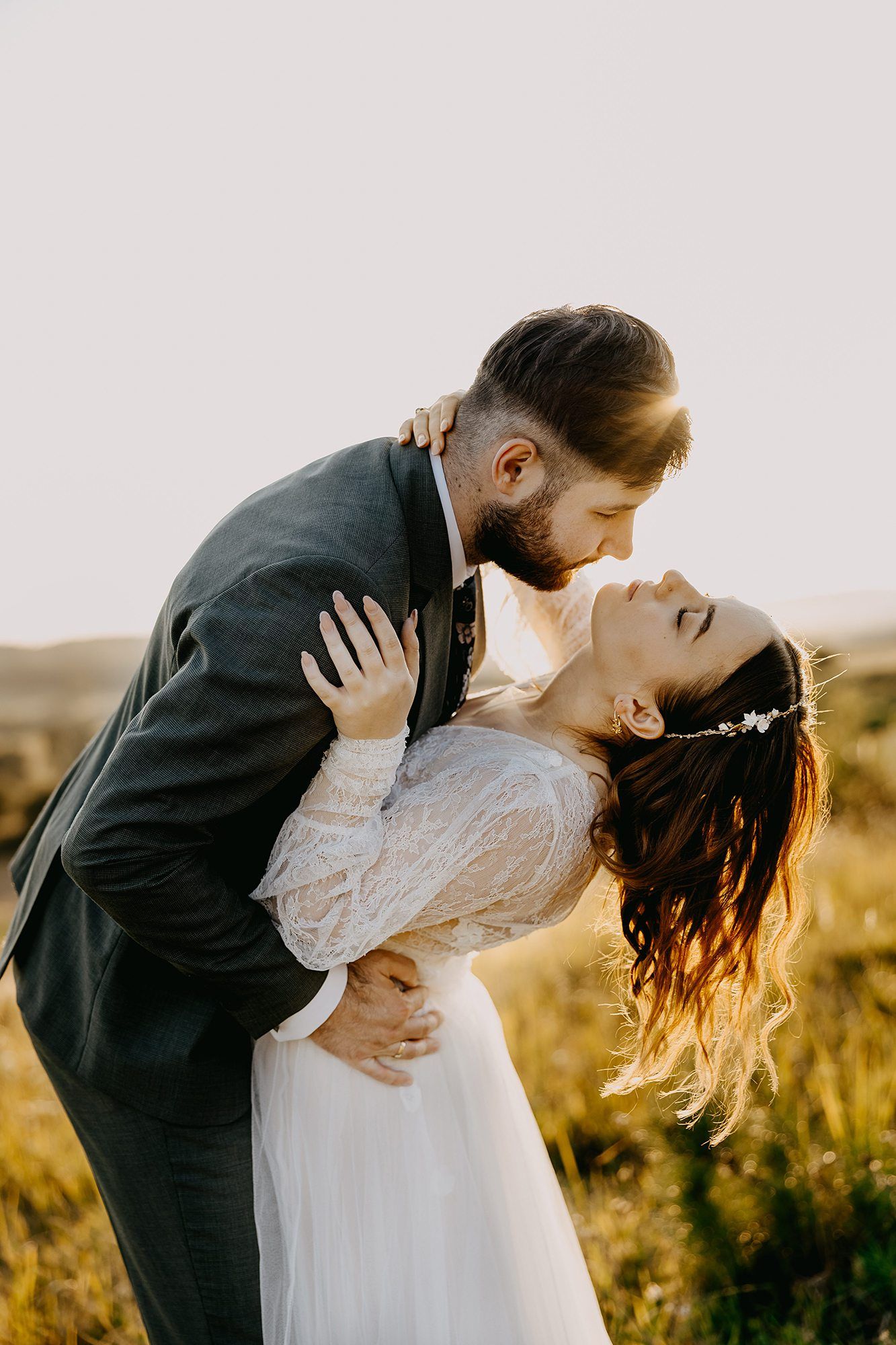 Groom dips bride for a kiss in golden sunlit field during romantic outdoor wedding photoshoot.