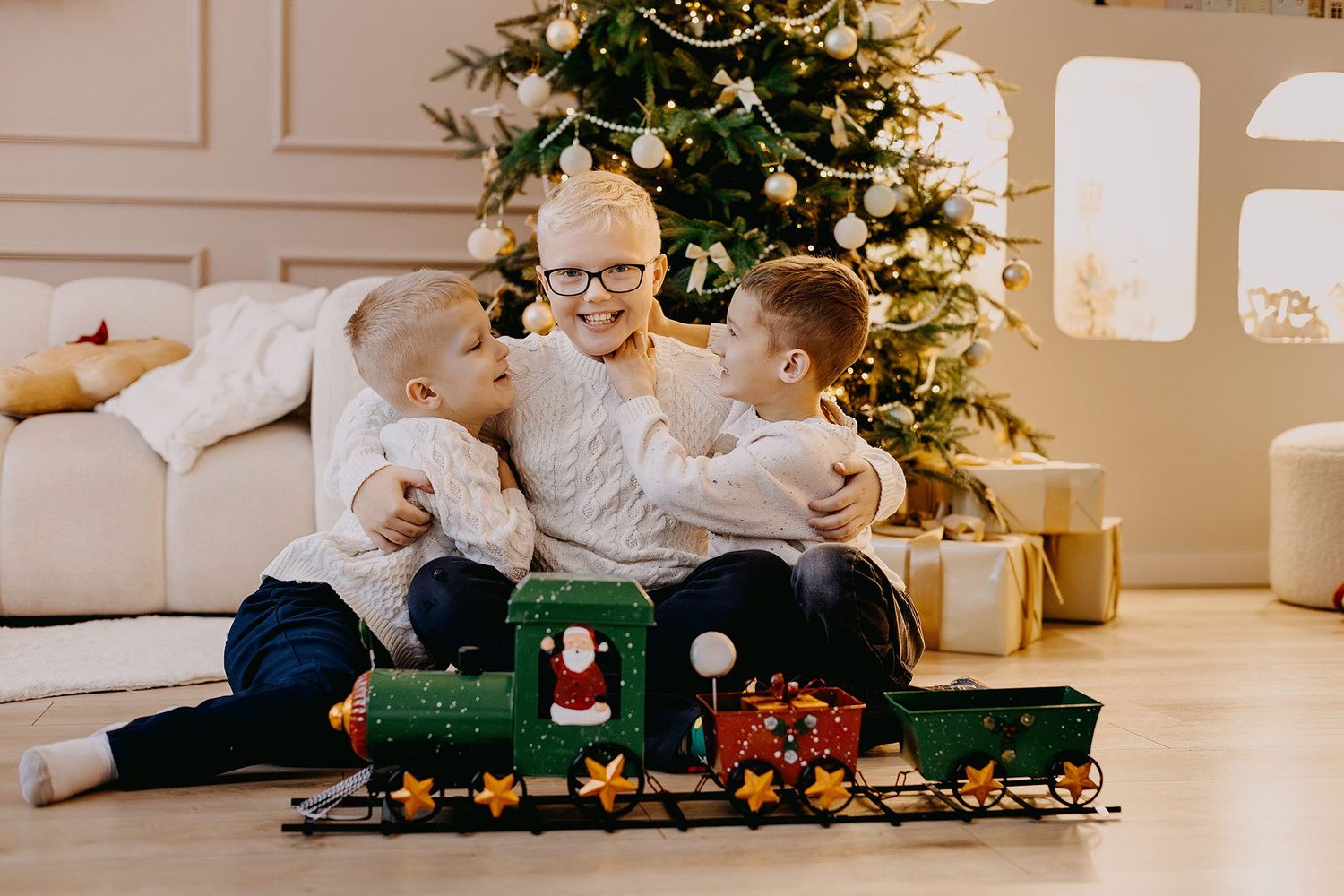 Three boys in white sweaters hugging and playing with a toy train set in front of a Christmas tree.