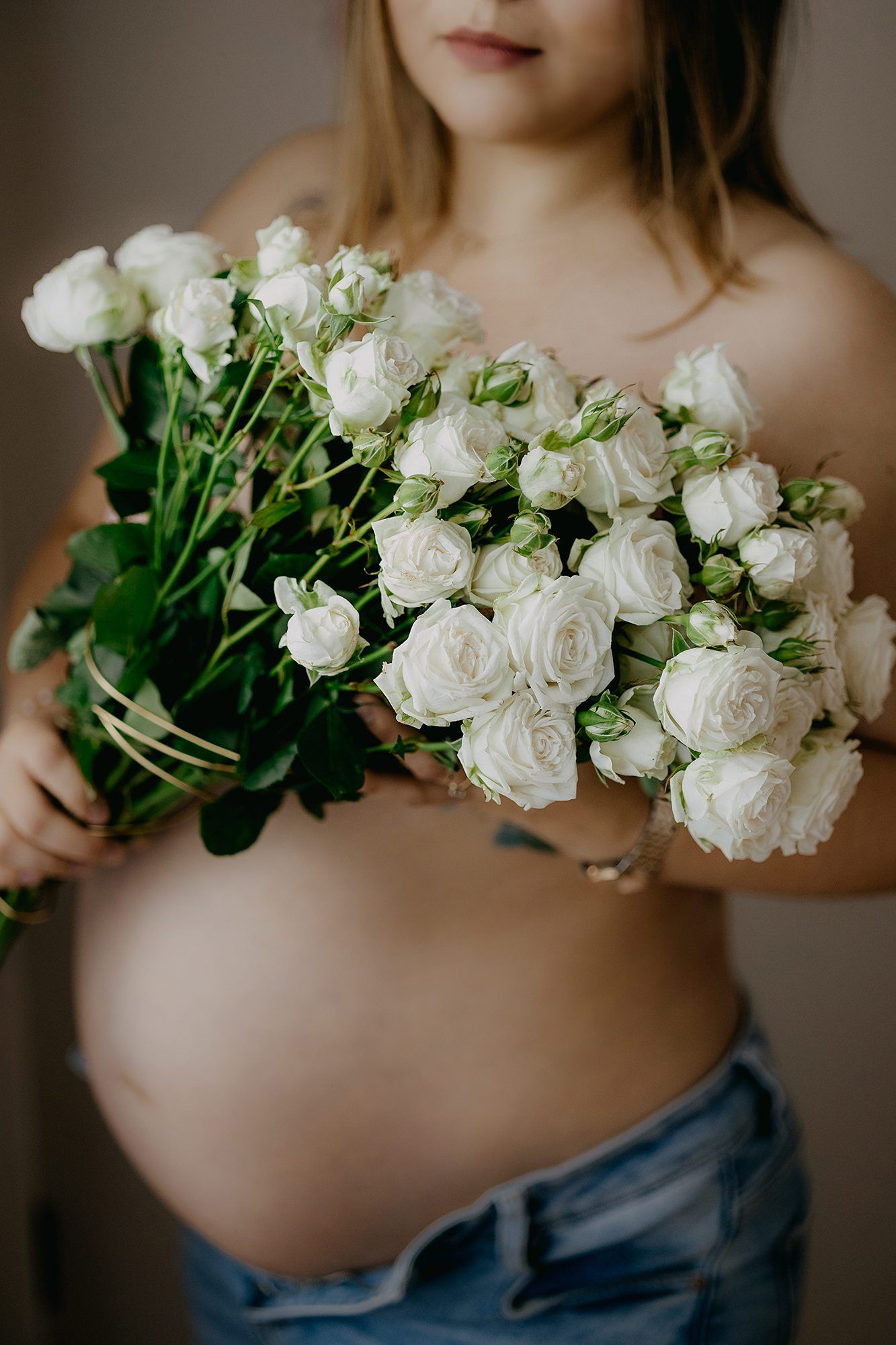 Pregnant woman holding a bouquet of white roses against her bare baby bump in a studio setting.