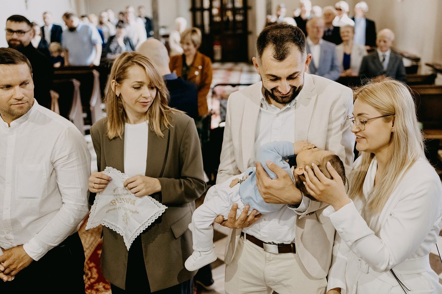 Group of elegantly dressed adults admire newborn baby during church baptism celebration.