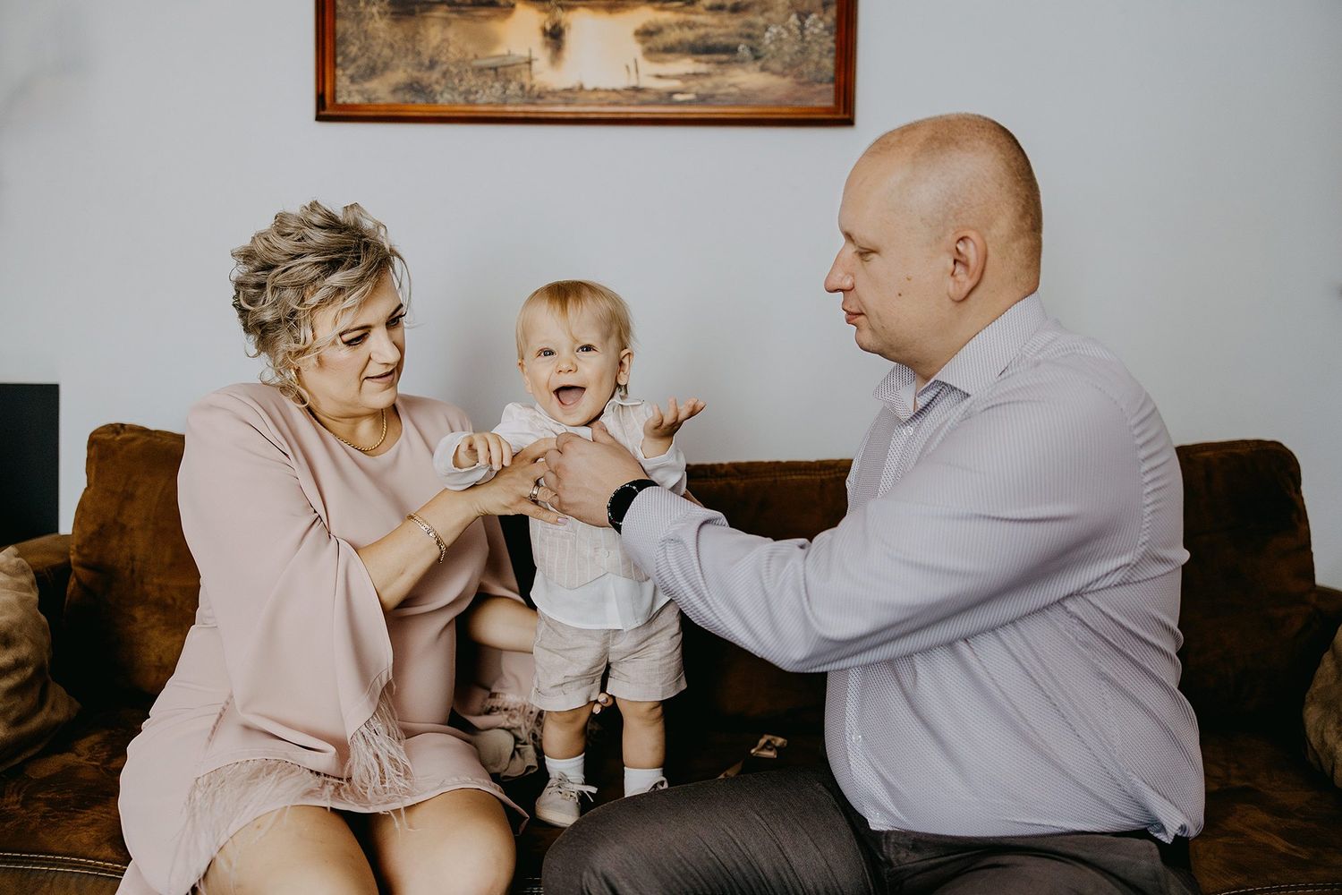 Grandparents sitting on a brown sofa helping their smiling toddler stand between them at home.