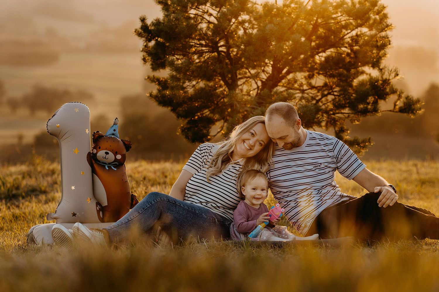 Family celebrating baby's first birthday outdoors at golden hour with number one balloon.