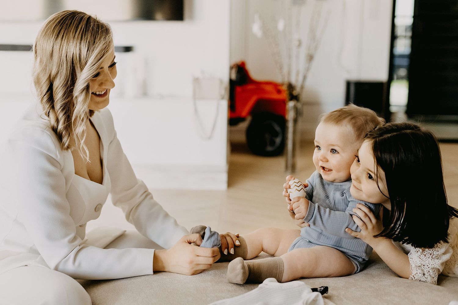A mother and older child play with a happy baby on a light living room floor.