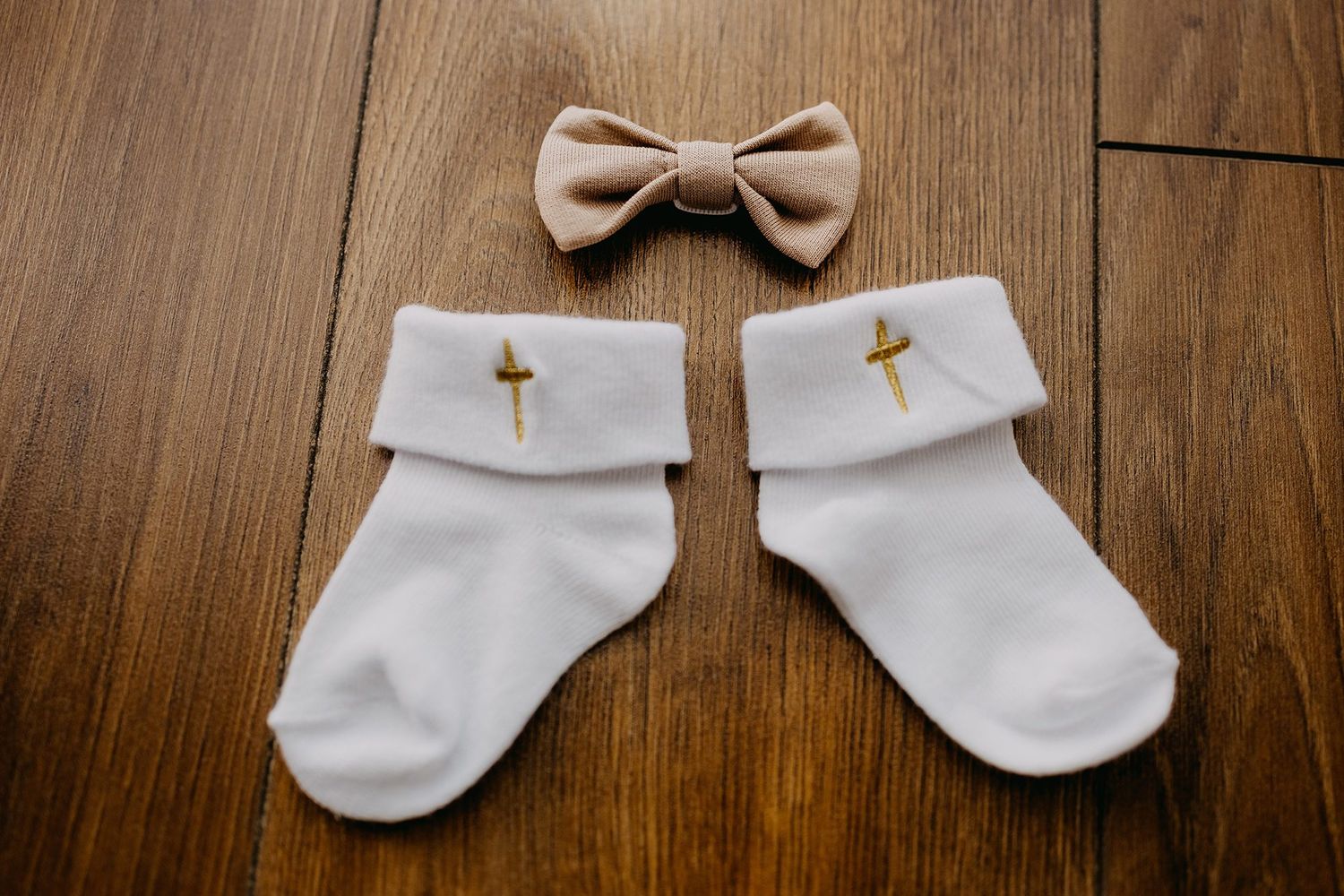 White baby baptism socks with gold cross embroidery and beige bow tie on wooden floor.