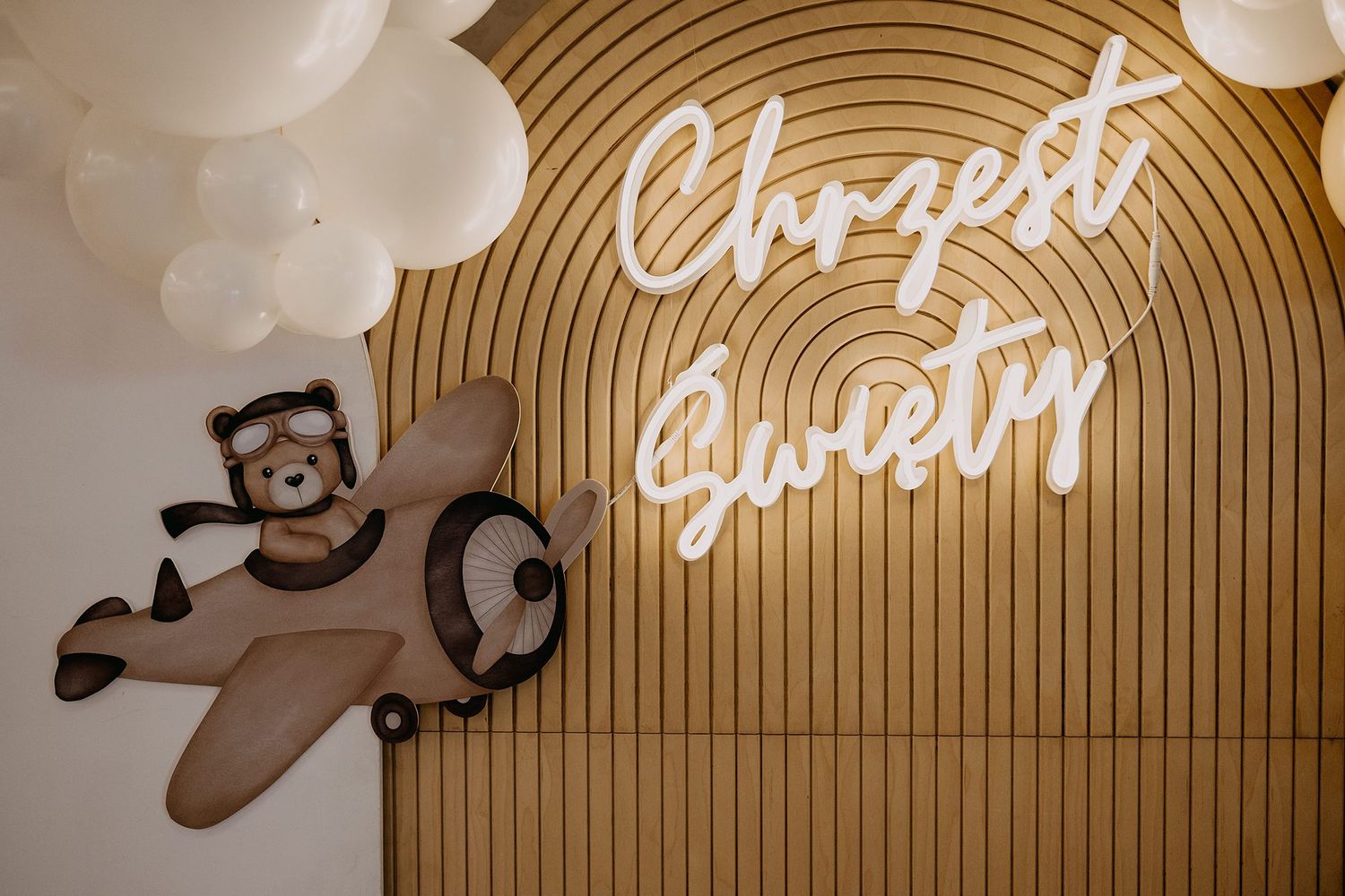 Decorative christening backdrop with neon Chrzest Swiety sign, teddy bear pilot, balloons, and wooden wall.