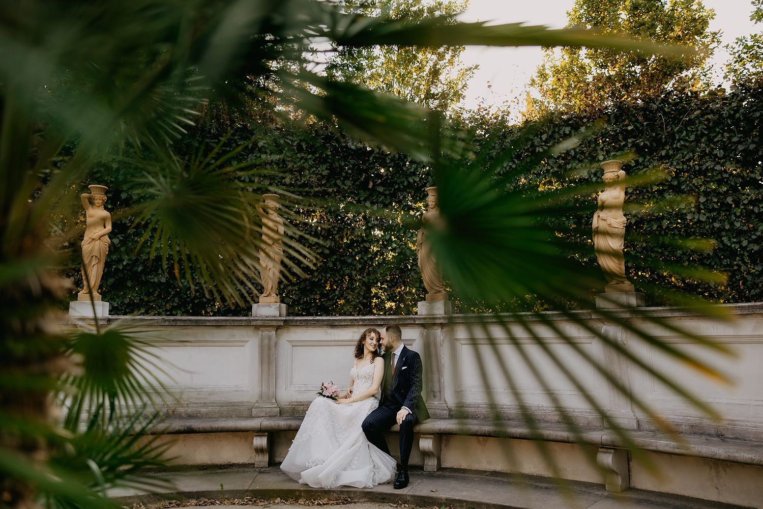 Bride and groom sitting together in an elegant garden with stone statues and lush greenery.