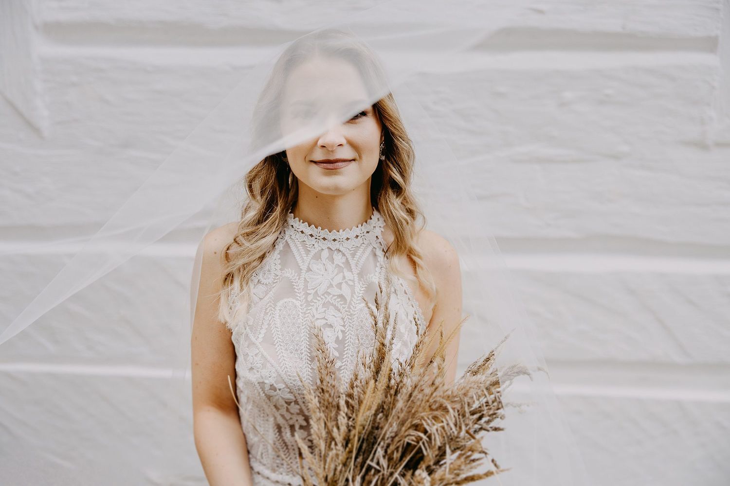 Smiling bride in lace gown holds dried pampas bouquet with veil draped over her face.