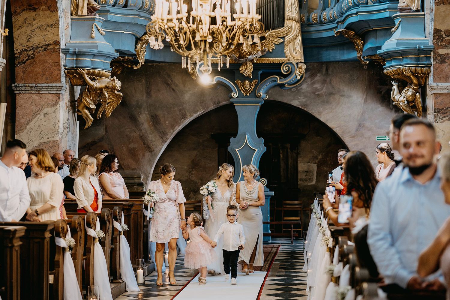 Wedding procession walking down the aisle of a stunning baroque church with guests watching.