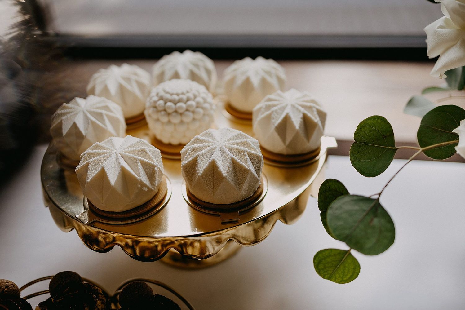 Elegant white fondant mini wedding cakes on a gold cake stand with greenery.