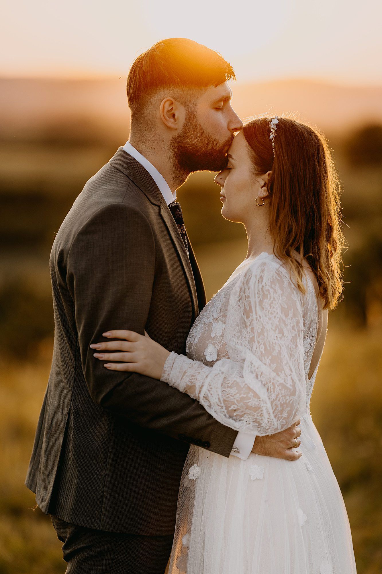Groom kissing bride on the forehead during a romantic sunset wedding photo in a field.