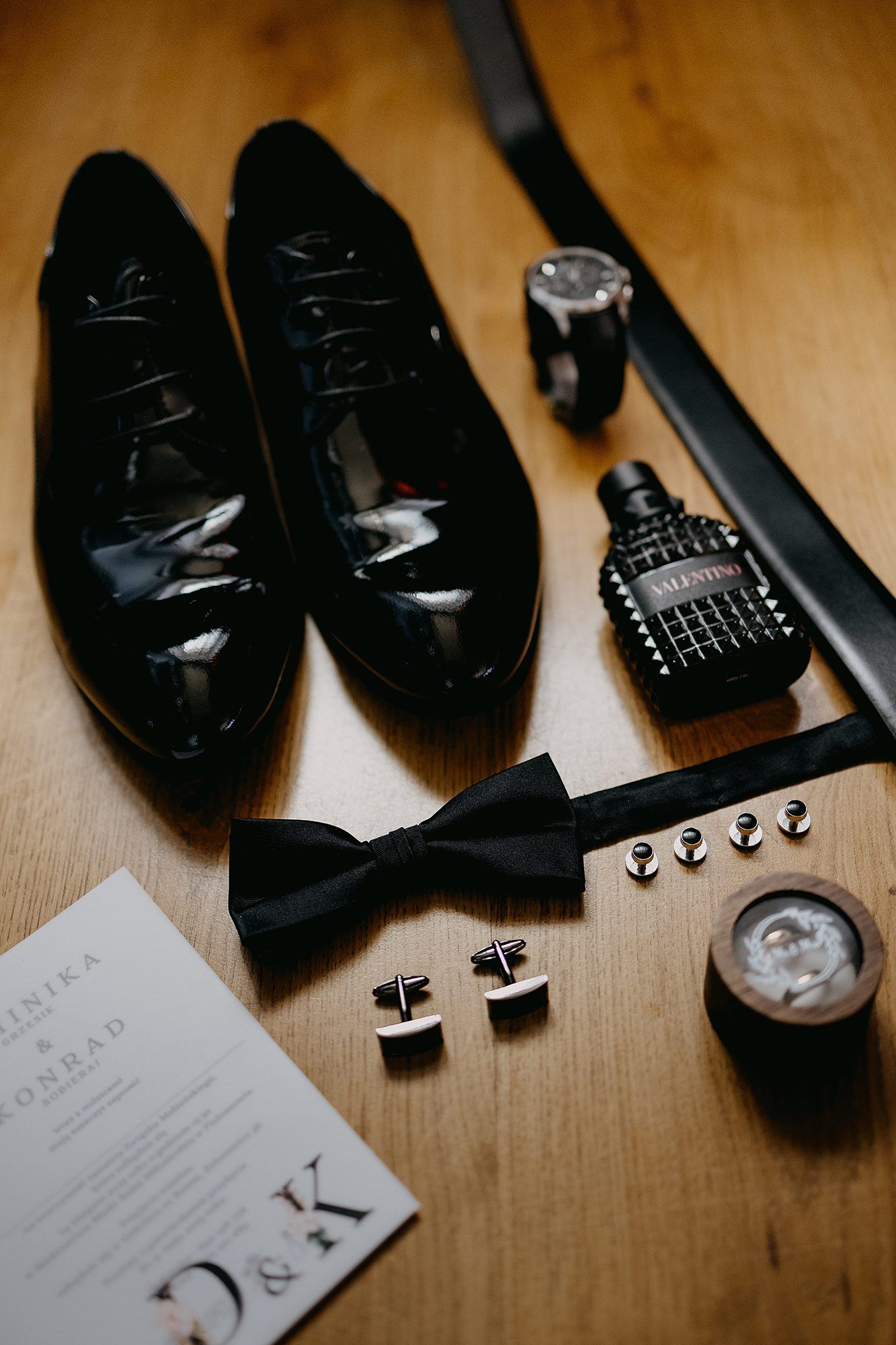 Groom wedding accessories flat lay featuring black patent shoes, bow tie, cufflinks, watch, belt, and Valentino cologne.