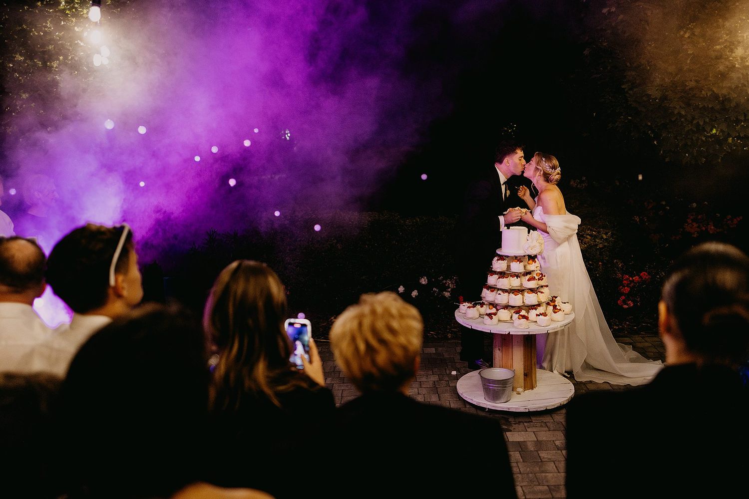 Bride and groom share a kiss beside their tiered wedding cake under dramatic purple smoke and fireworks.