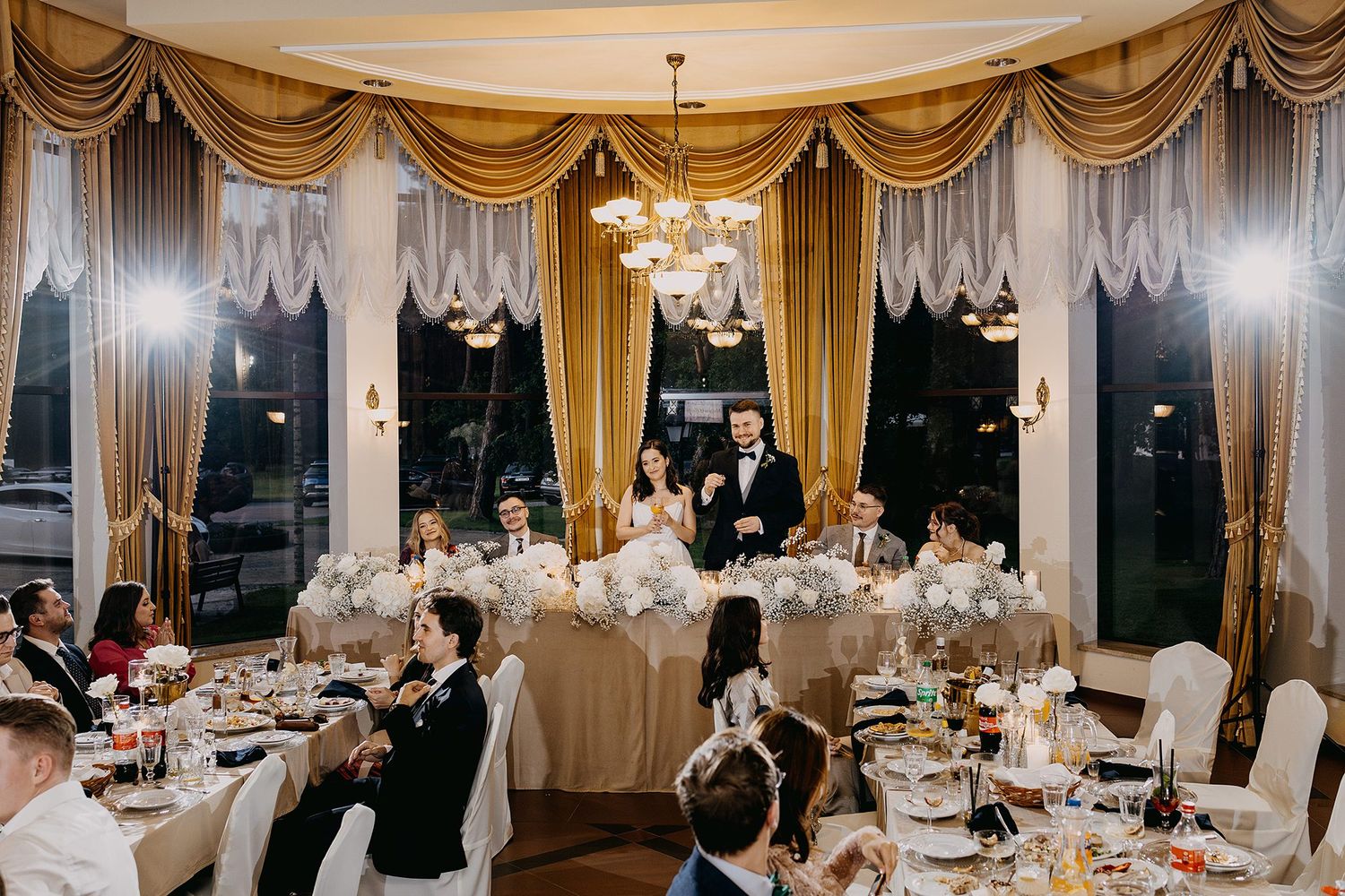 Bride and groom at elegant wedding reception with guests seated at decorated tables.