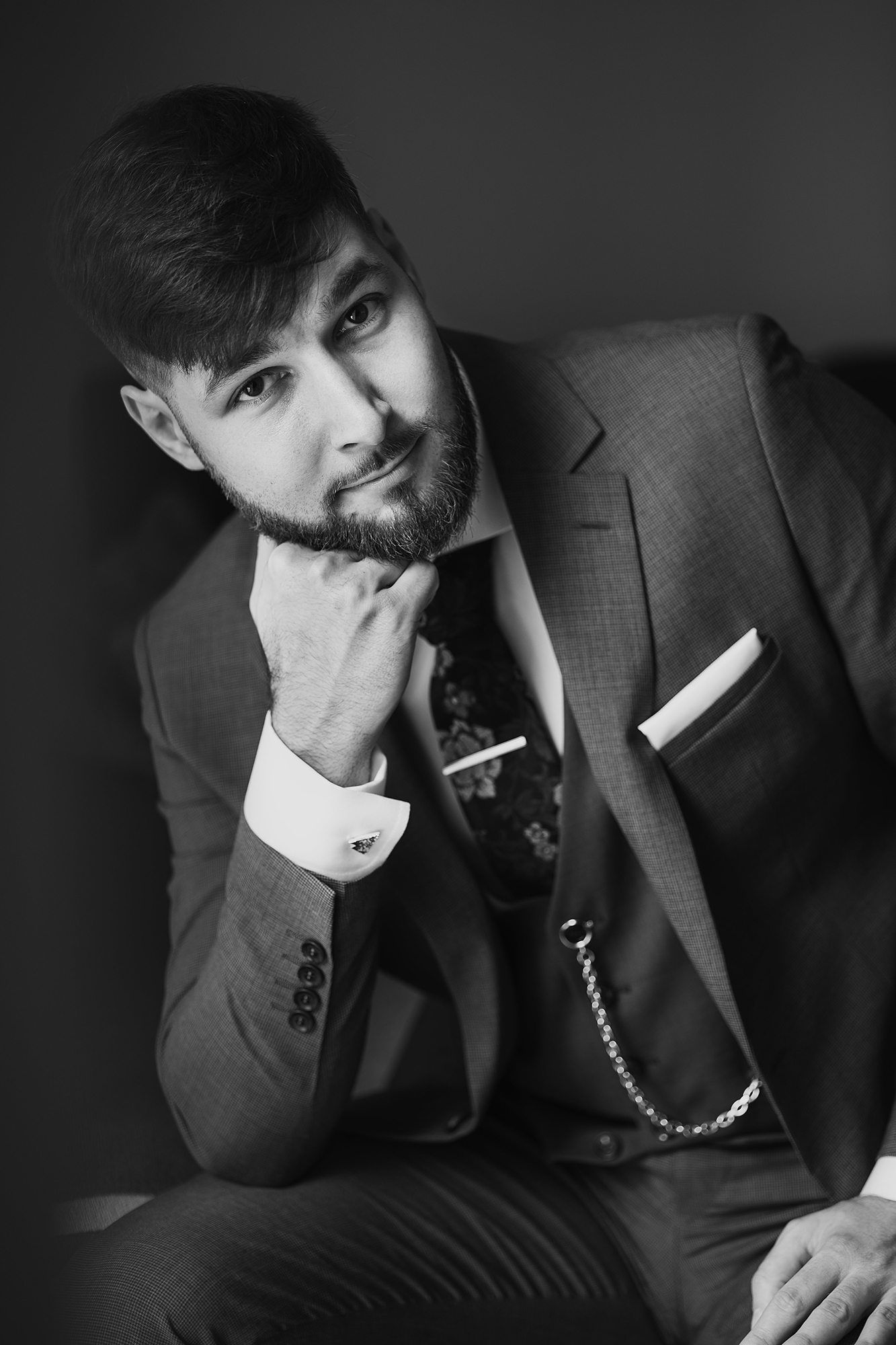 Elegant man in suit with pocket square and chain, posing thoughtfully in black and white portrait photo.