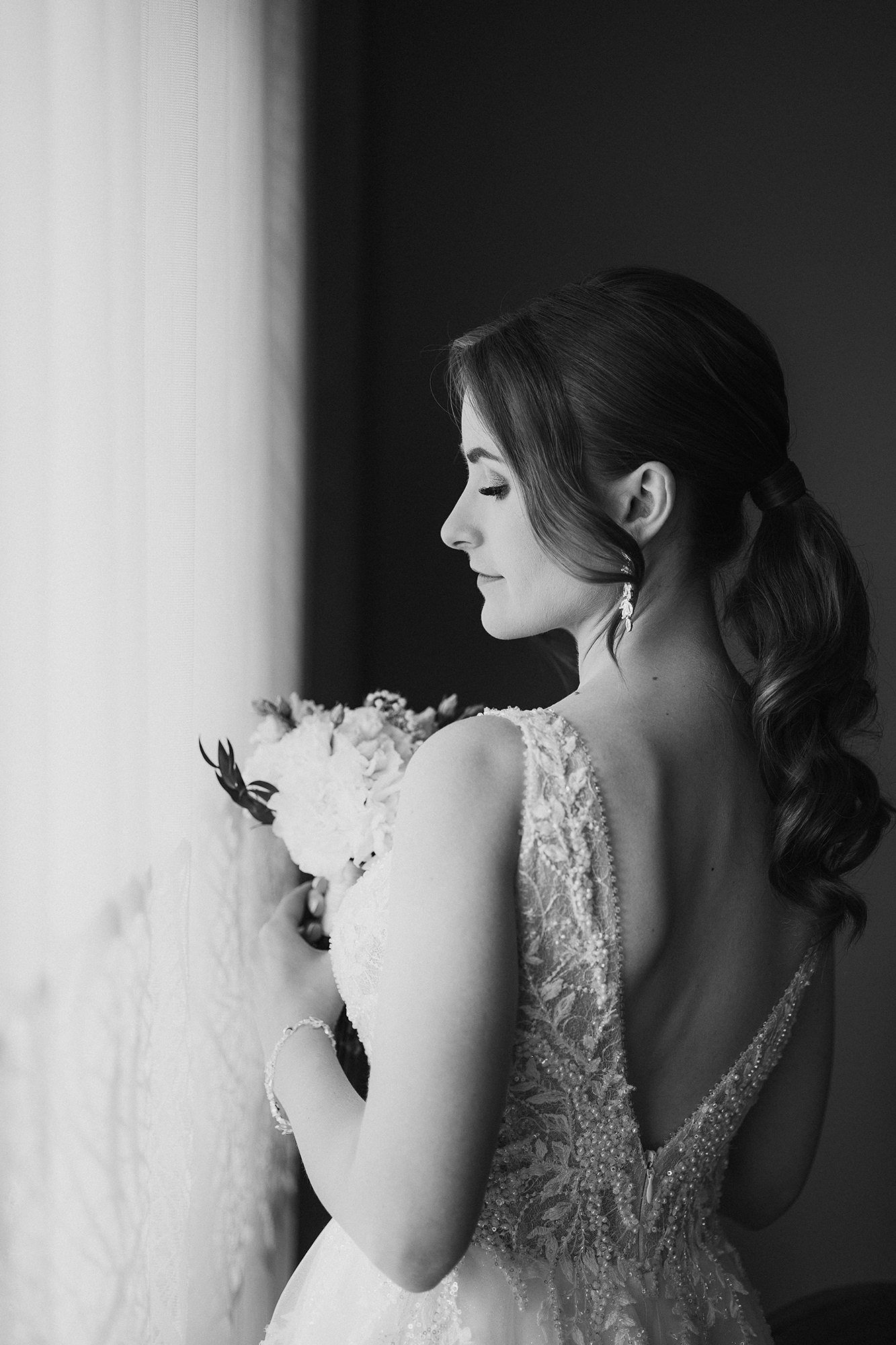 Black and white portrait of bride holding bouquet while gazing out window in lace gown.