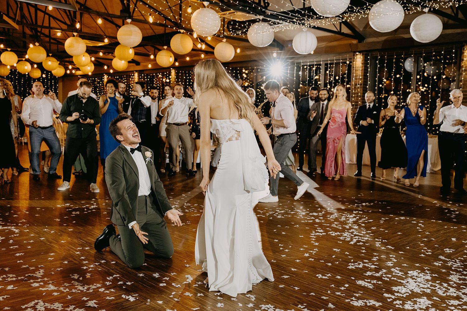 Groom kneeling before bride on dance floor surrounded by guests and paper lanterns.