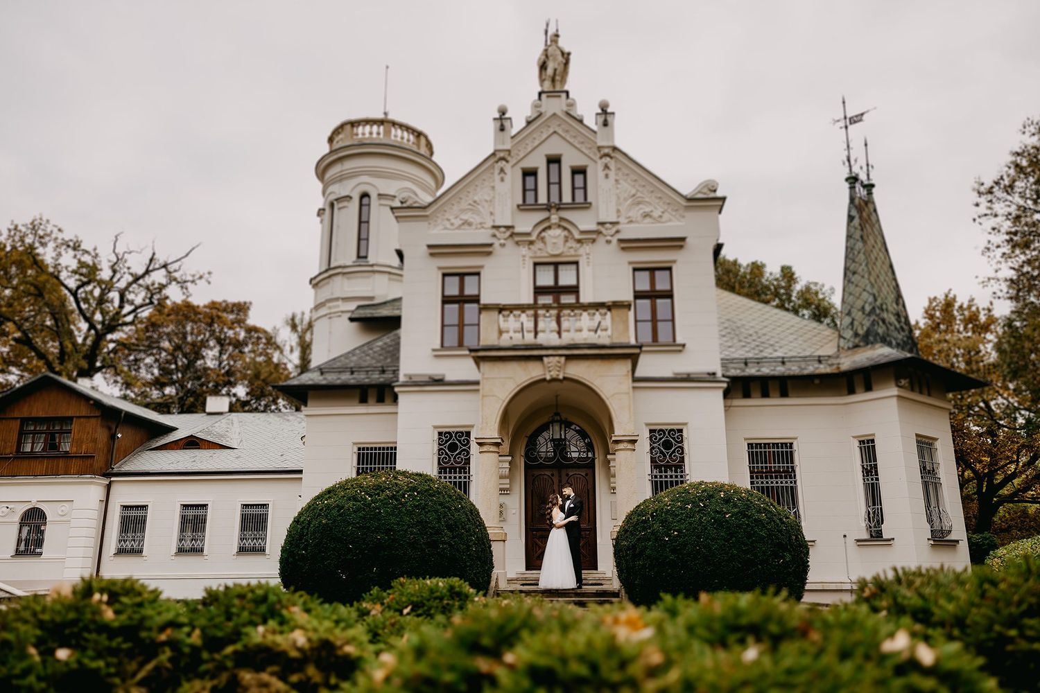 Elegant wedding couple poses before a grand white historic manor surrounded by manicured hedges.