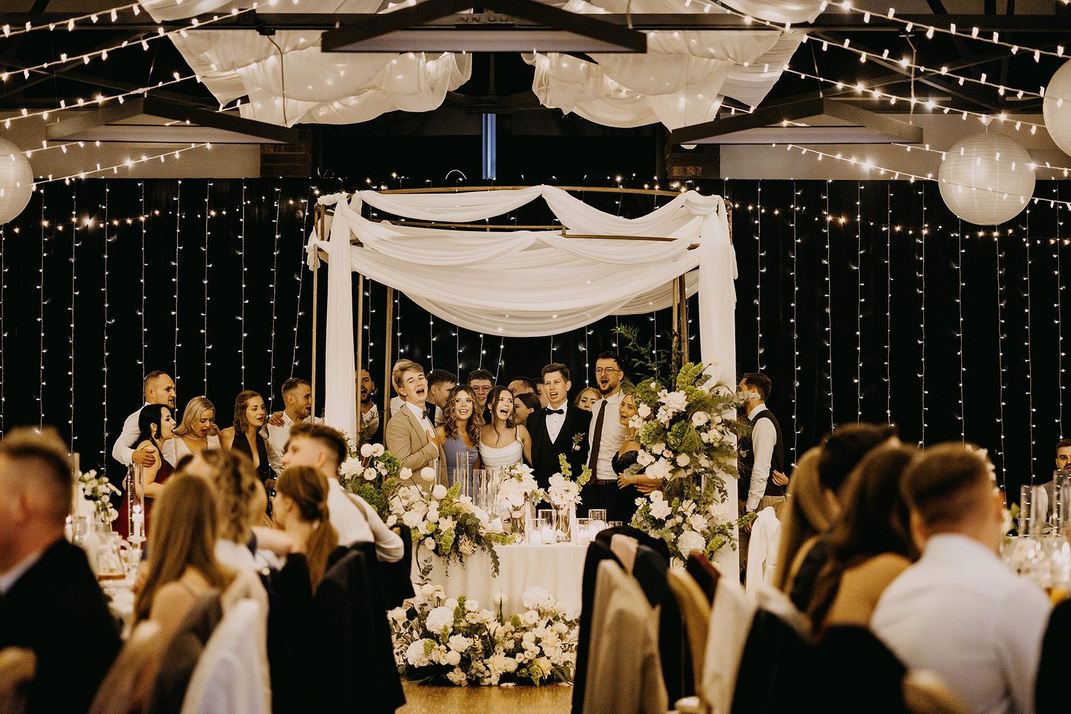 Elegant wedding reception with fairy lights, floral arrangements, and wedding party gathered under a draped canopy.