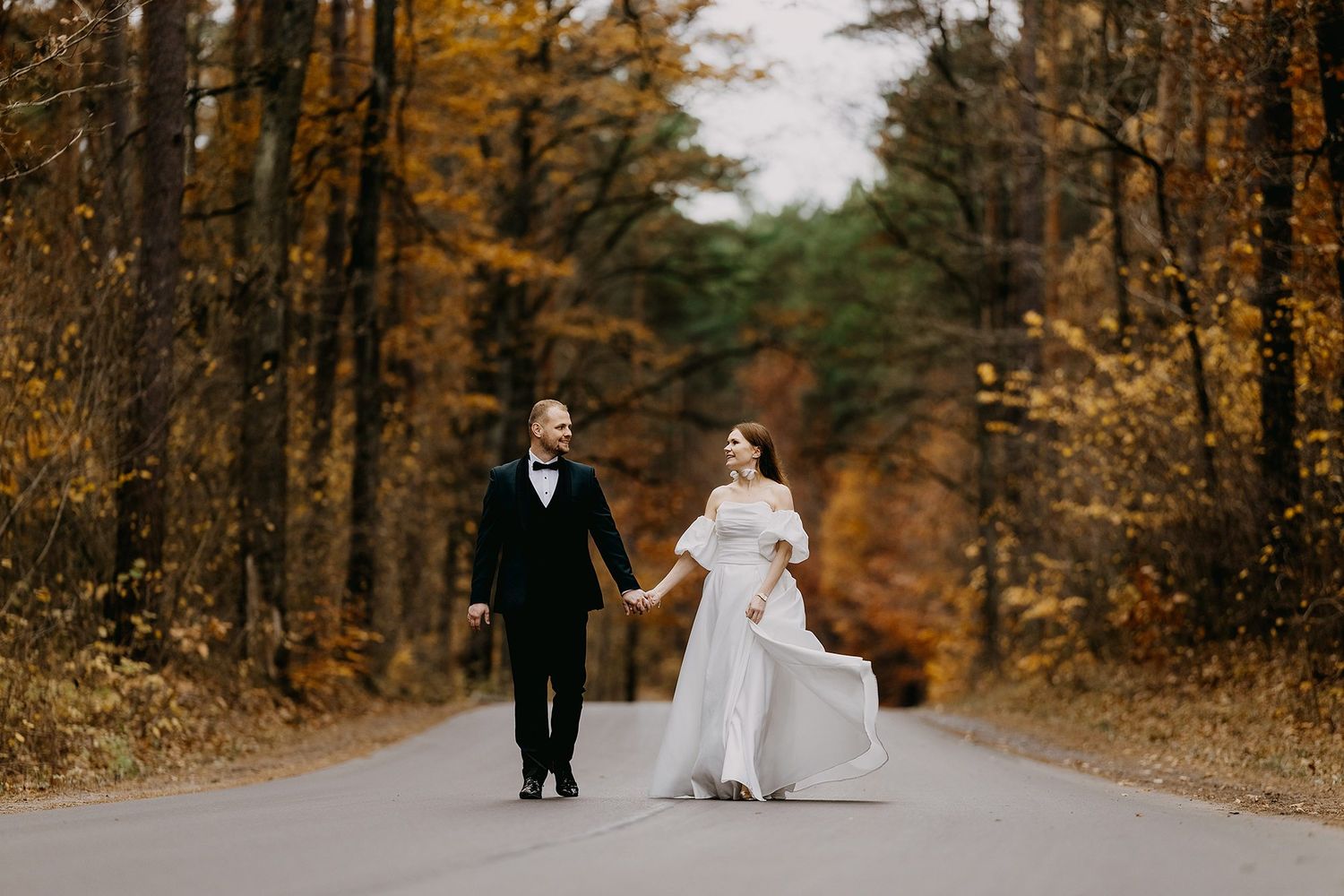 Bride and groom walk hand-in-hand down a tree-lined road surrounded by vibrant autumn foliage.
