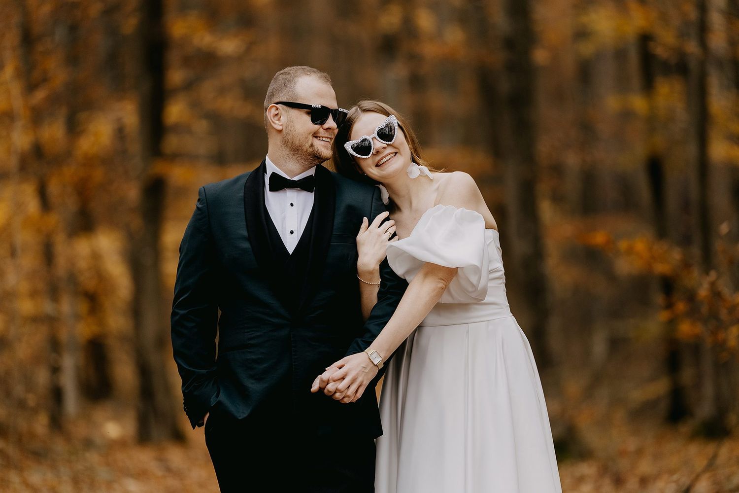 Stylish bride and groom wearing sunglasses holding hands in an autumn forest with golden foliage.