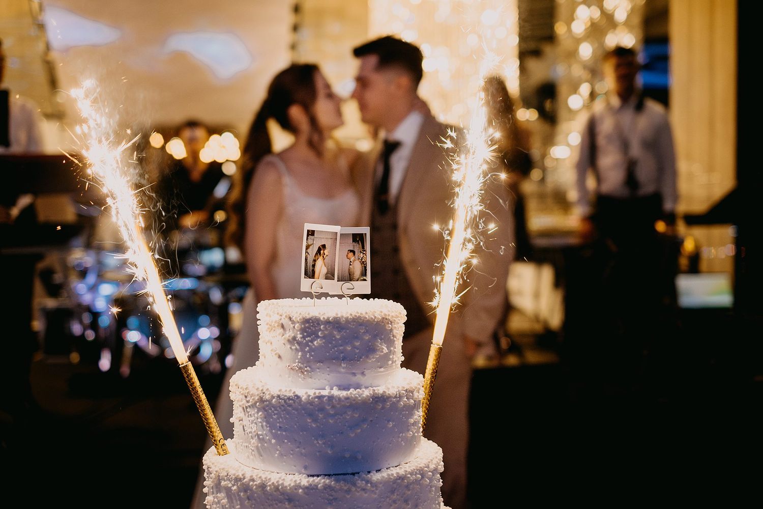 Three-tier white wedding cake with sparklers as bride and groom share a kiss in the background.