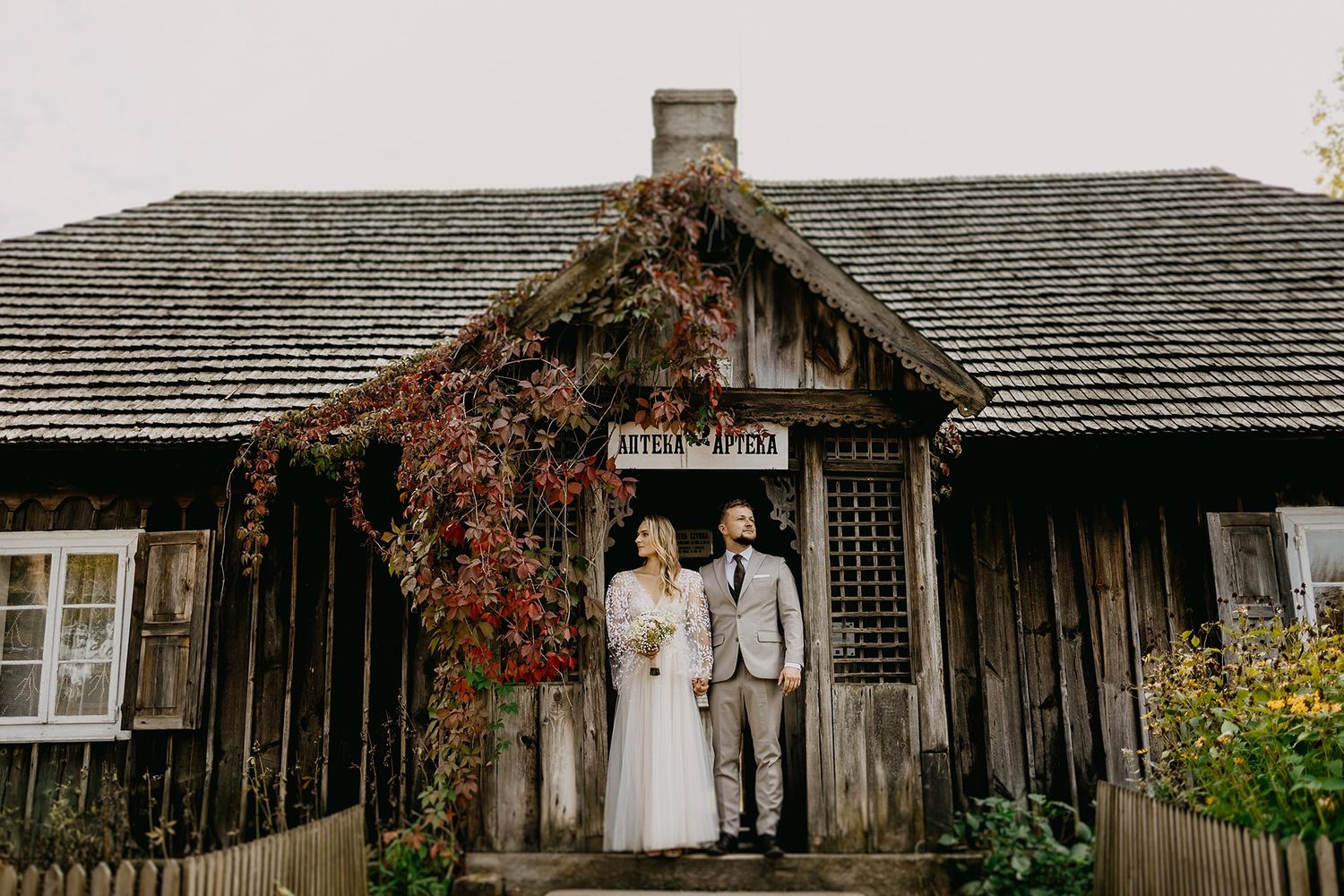 Bride and groom stand before a rustic wooden building covered in red ivy on their wedding day.