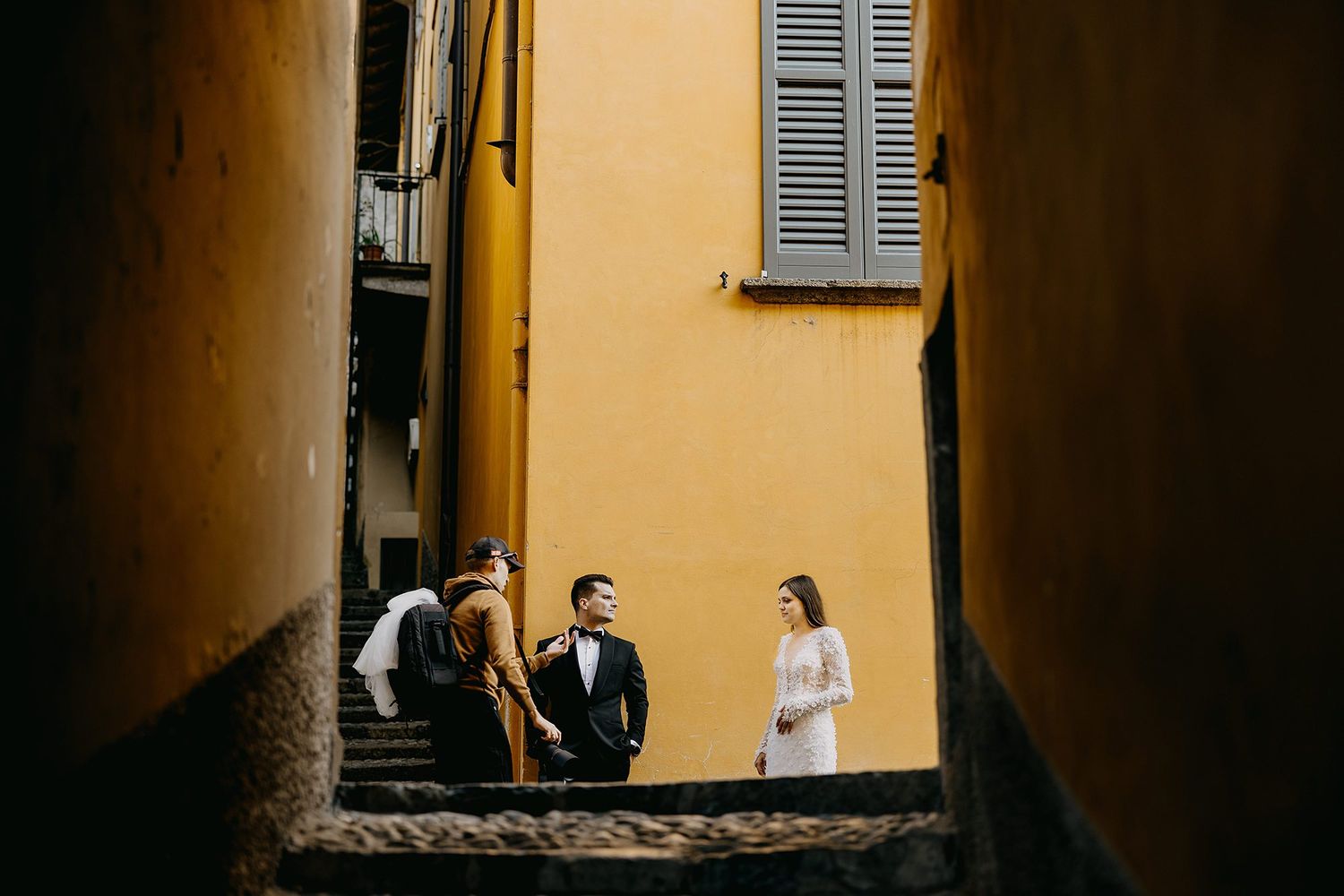Wedding photographer capturing bride and groom on narrow cobblestone stairs between yellow walls.