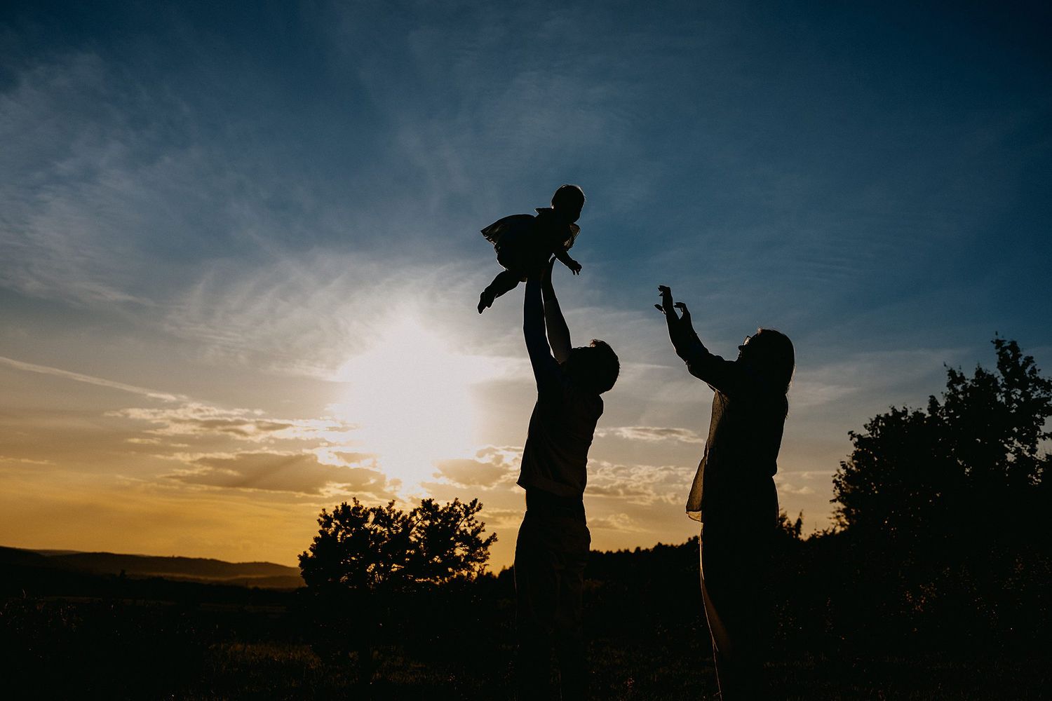 Silhouette of parents lifting baby at sunset with golden sky and trees in background.