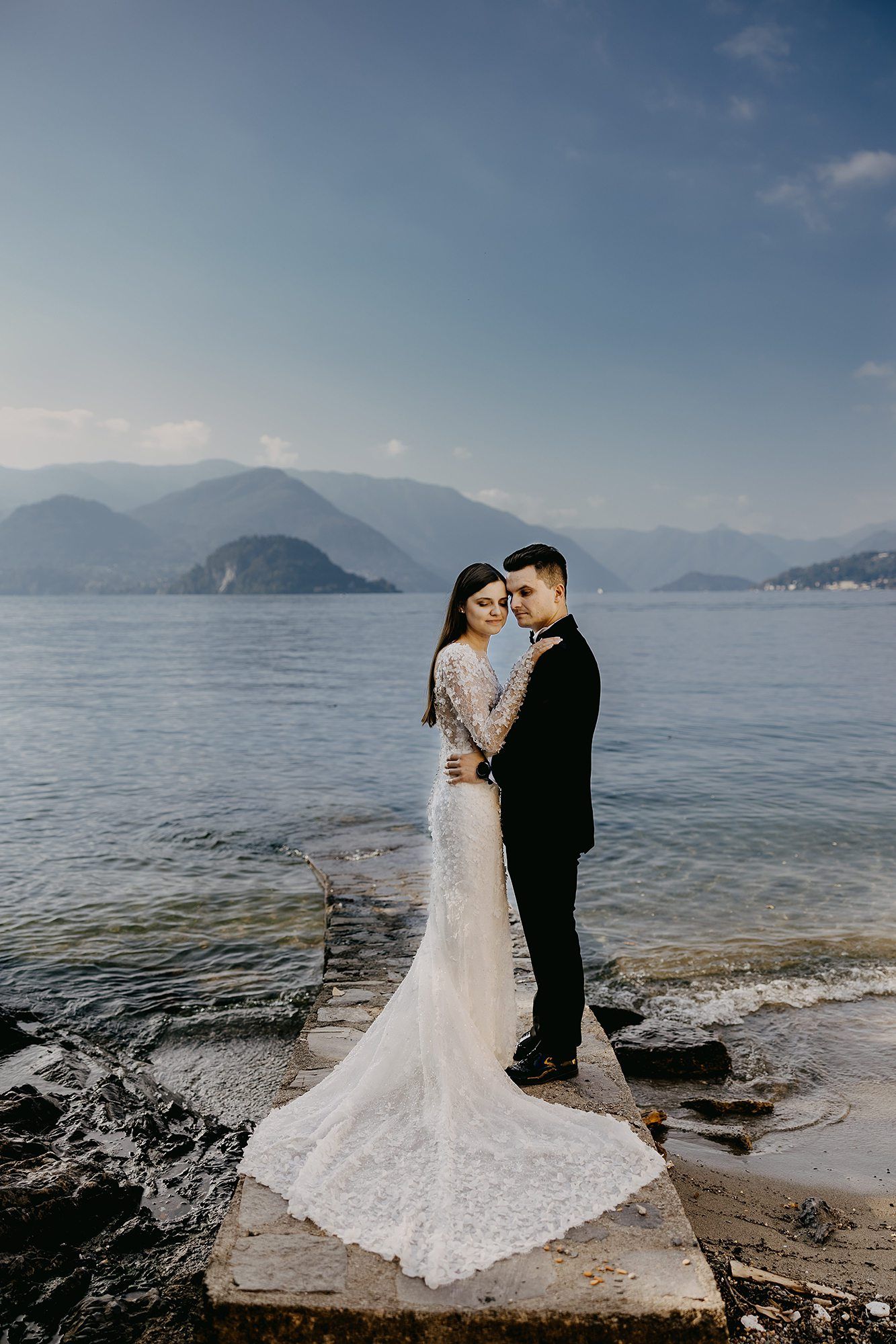 Wedding couple embraces on rocky lakeside with stunning mountain backdrop and serene blue waters.