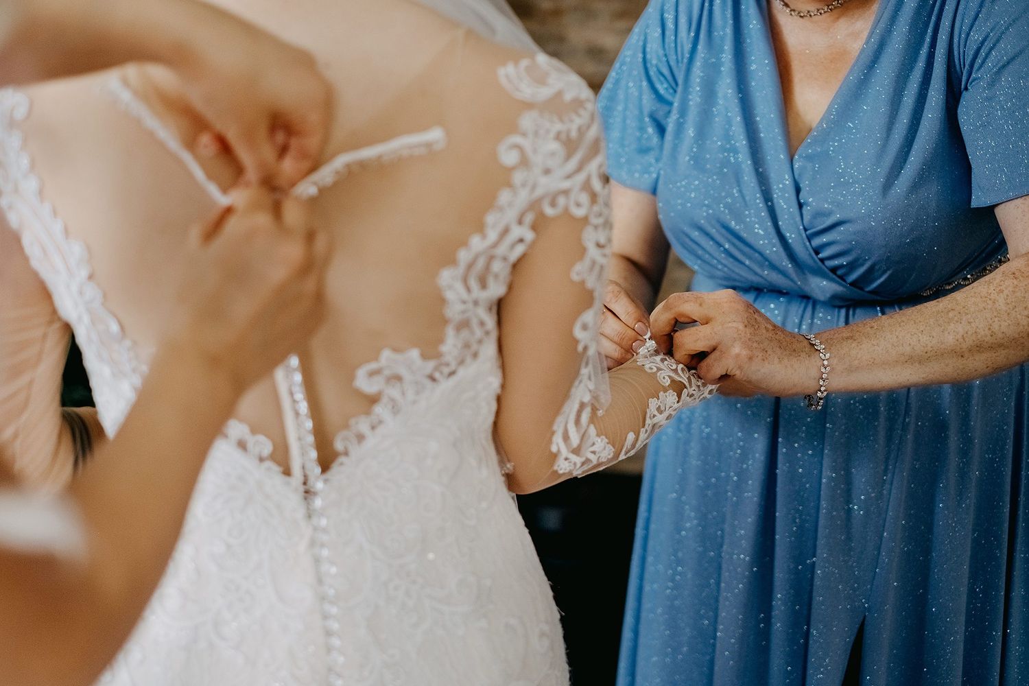 Mother helping bride button intricate lace back detail of wedding gown during bridal preparations.