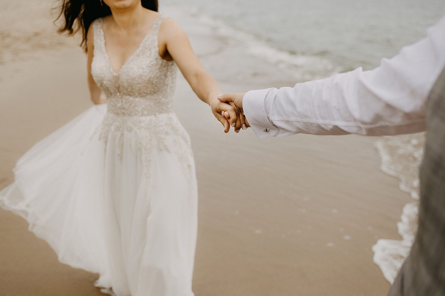 Bride in beaded wedding gown holds groom's hand while walking along ocean shoreline.