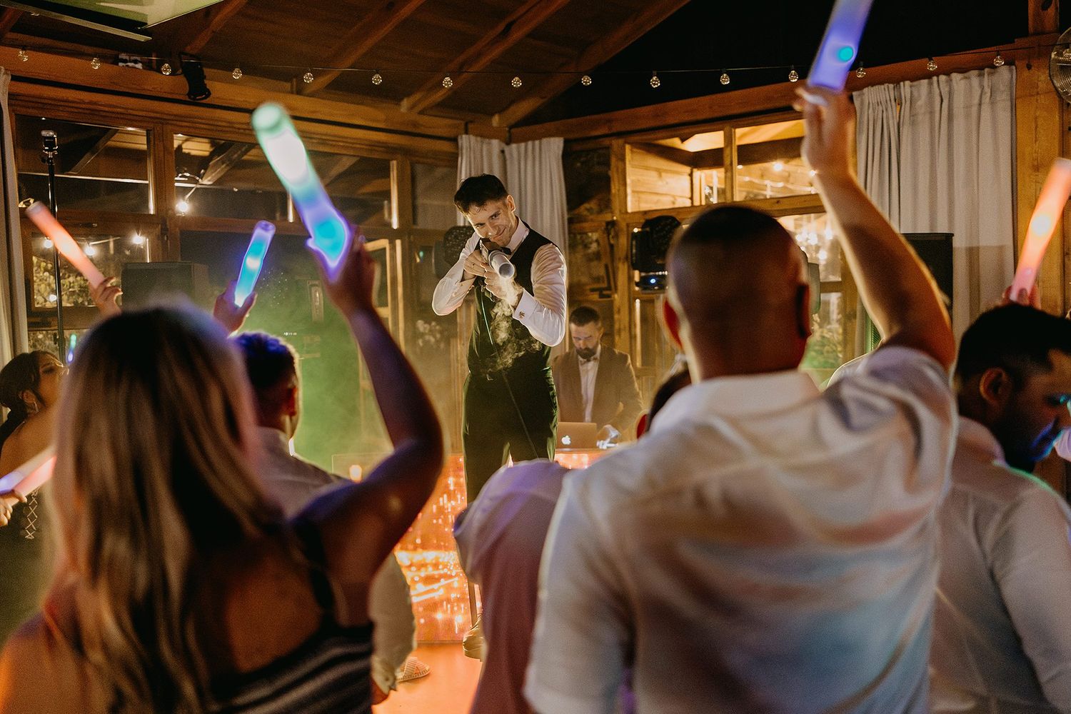 Energetic wedding reception party with guests raising glow sticks toward a performing entertainer on stage.