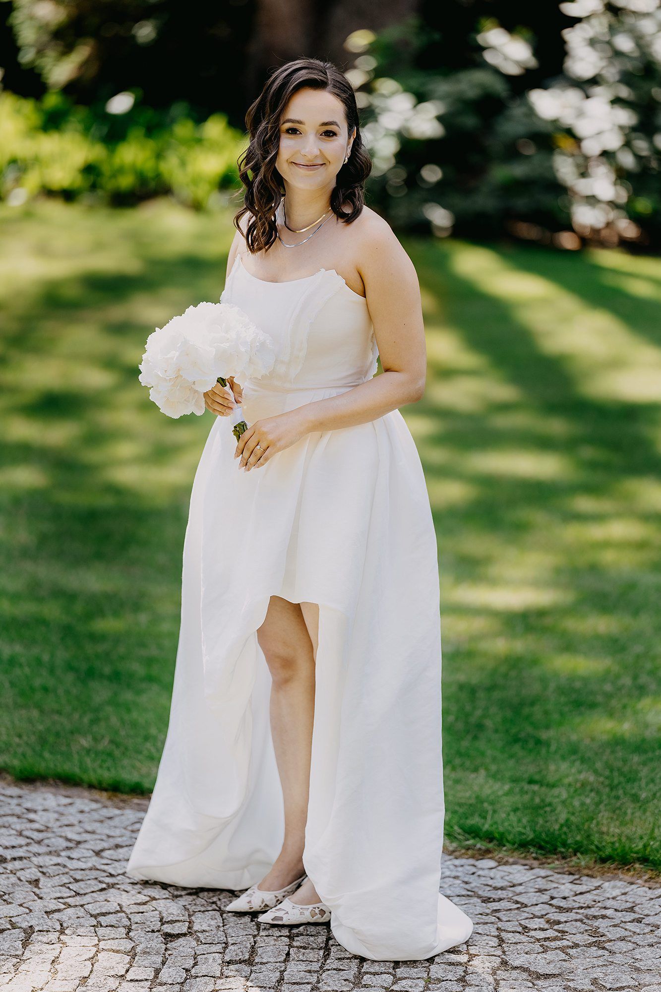 Radiant bride in strapless white high-low gown holding white peony bouquet in sunlit garden.