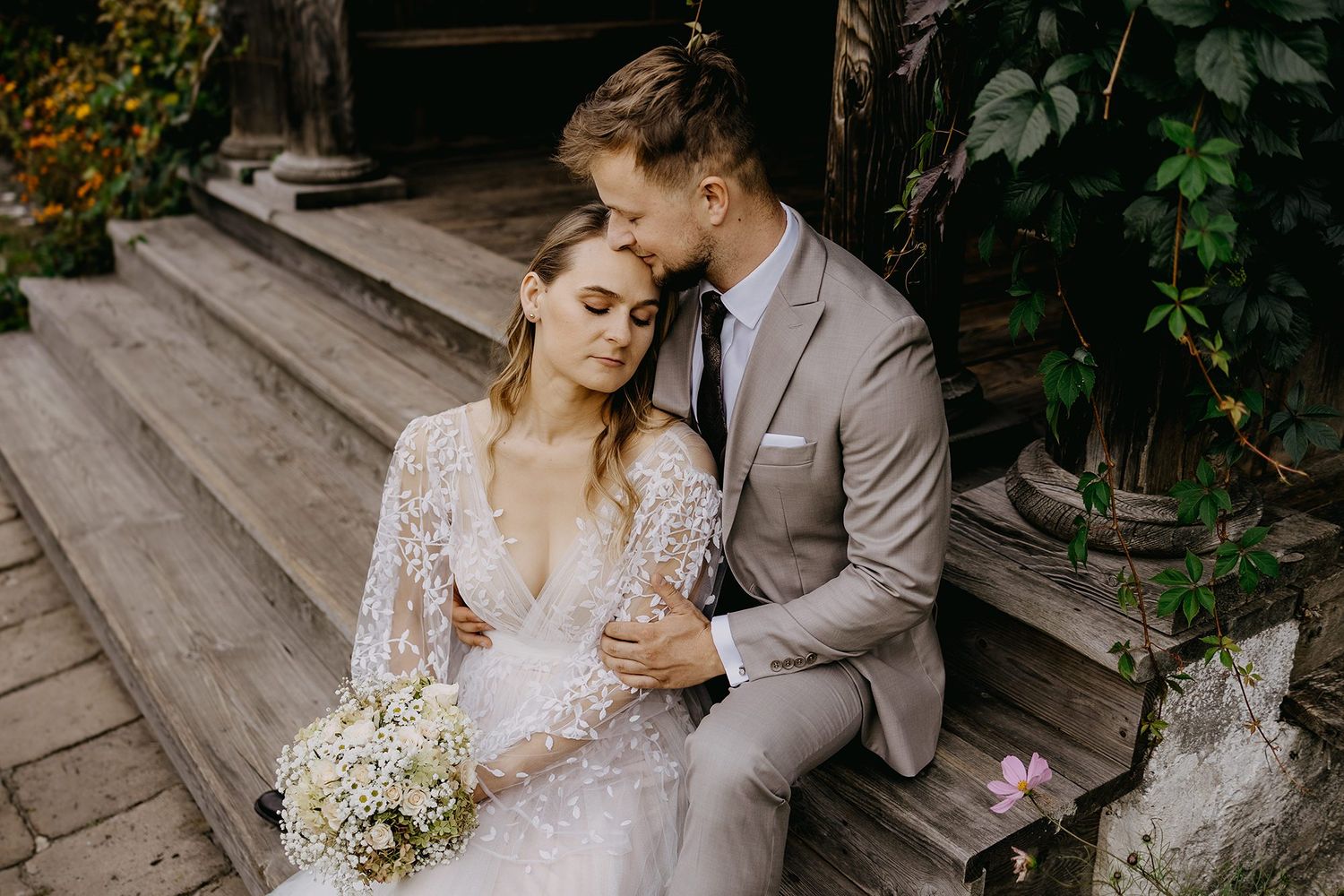 Bride with bouquet and groom sitting on wooden steps in garden.