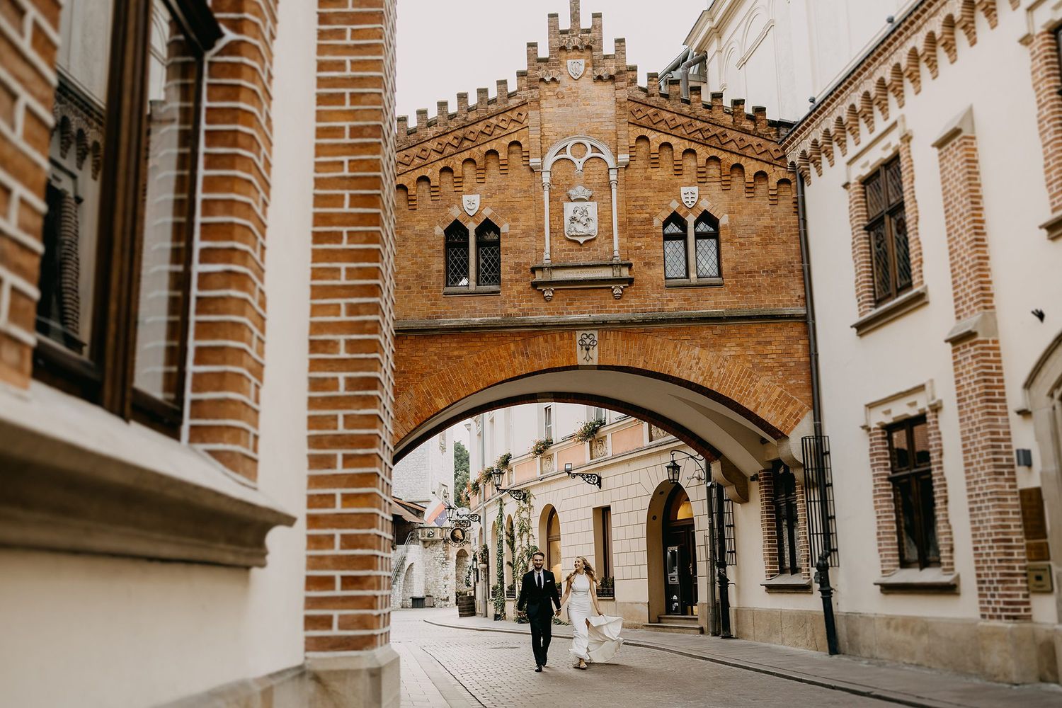 Wedding couple strolling beneath historic brick arch bridge on charming European street.